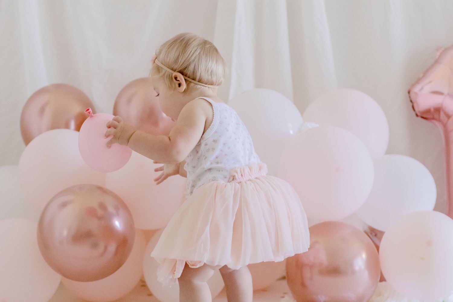 Toddler in white and pink dress climbing over pink and rose gold balloon arrangement.