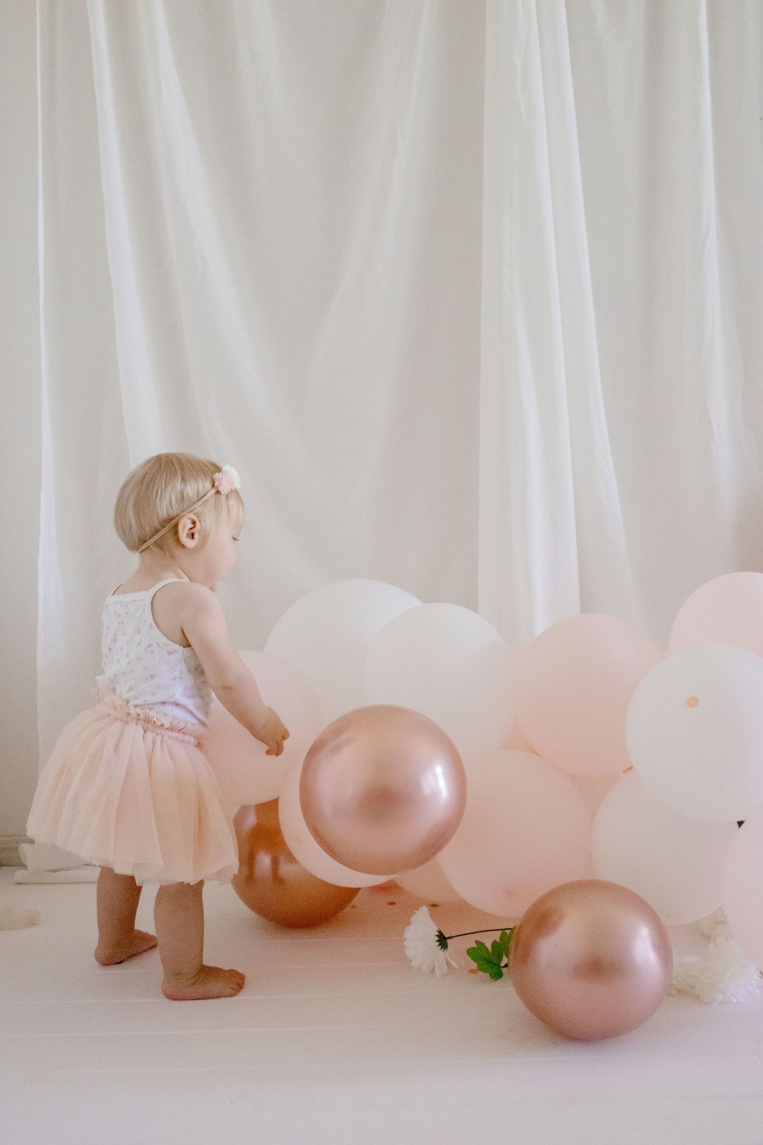Little one in pink tutu dress exploring balloon decorations against white backdrop.