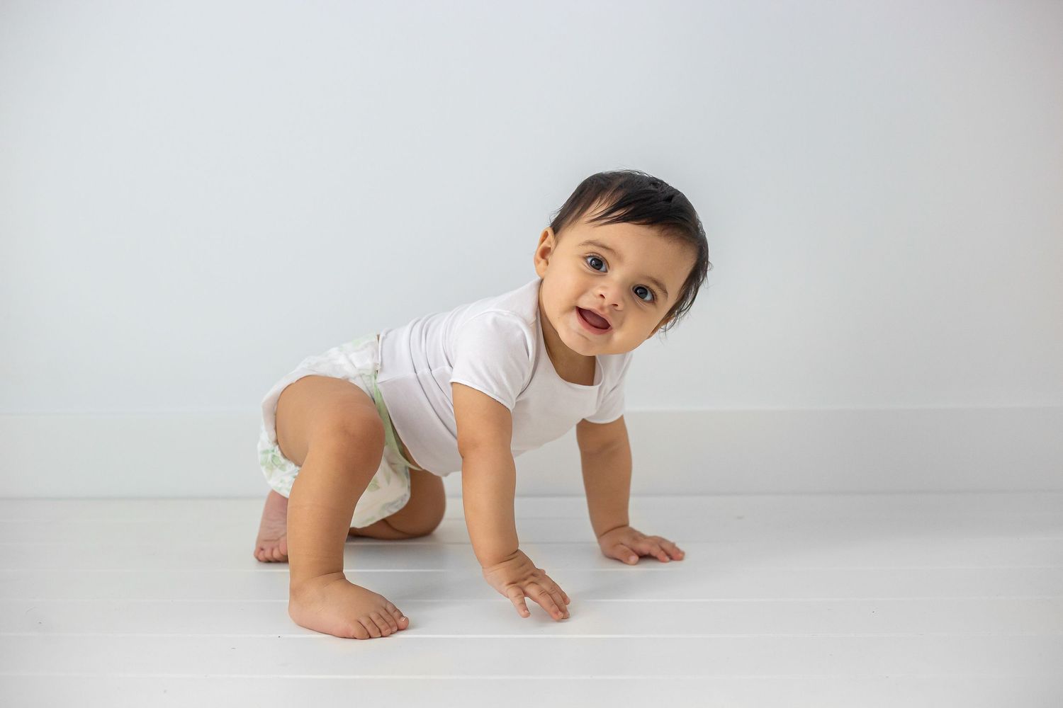 Baby wearing white onesie crawls on white floor while smiling joyfully during studio photoshoot.