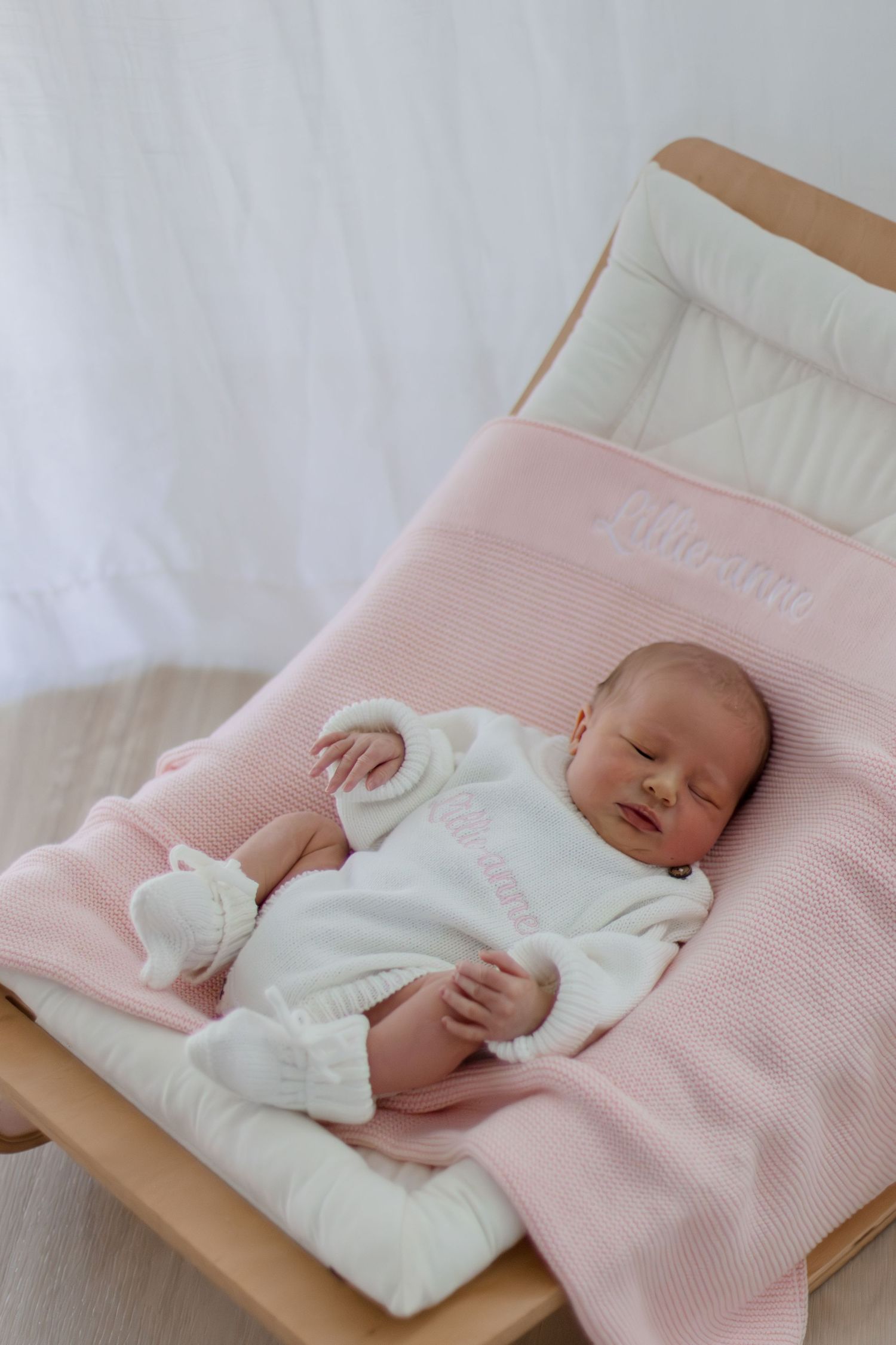 Newborn baby sleeping peacefully on a pink knit blanket in a wooden bouncer chair.