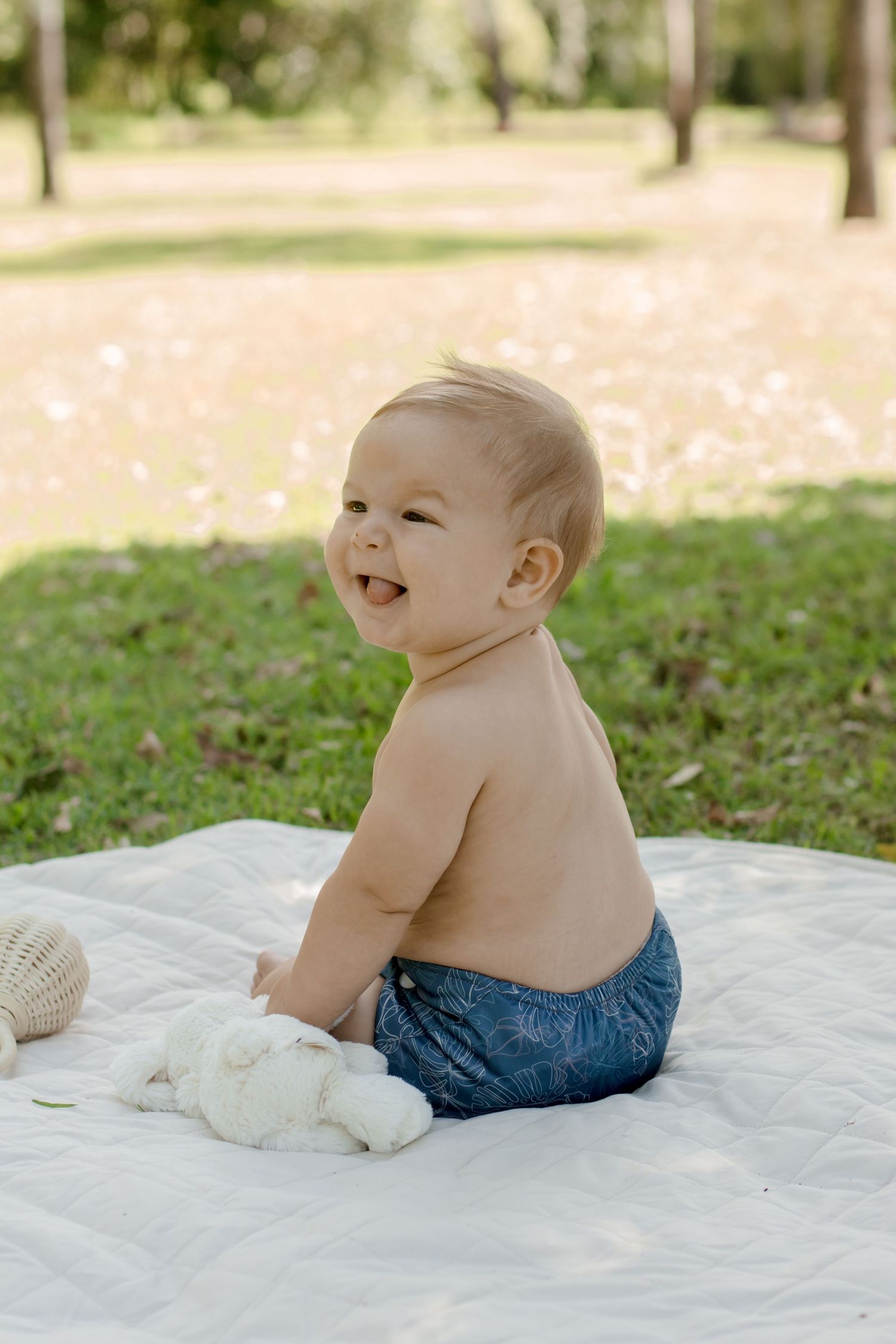 A happy baby wearing blue shorts sits on a white blanket outdoors in a sunny garden setting.