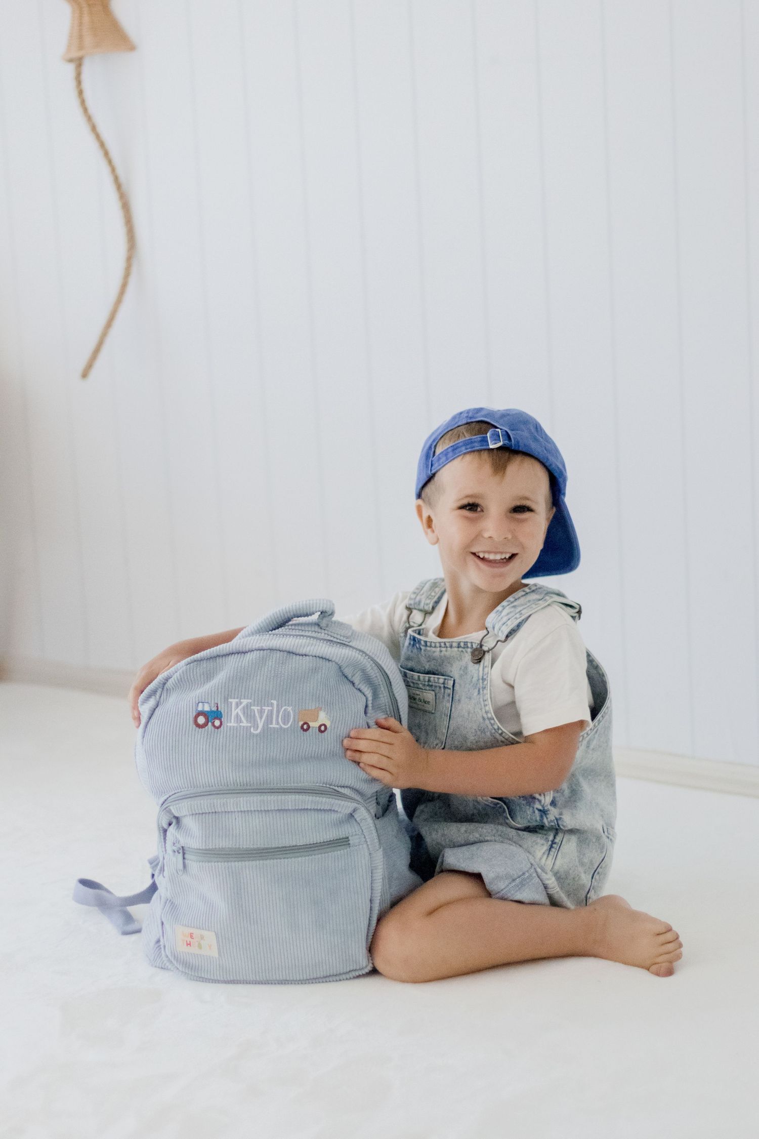 Young child sitting cross-legged with a light blue backpack in a bright white room.