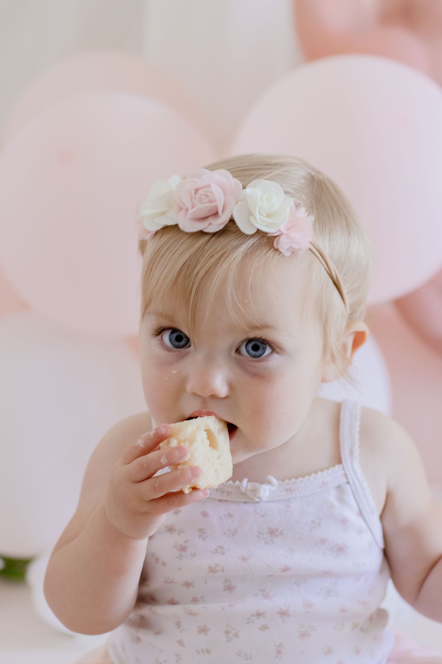 Baby in white dress with rose headband eating snack with pink balloons in background.