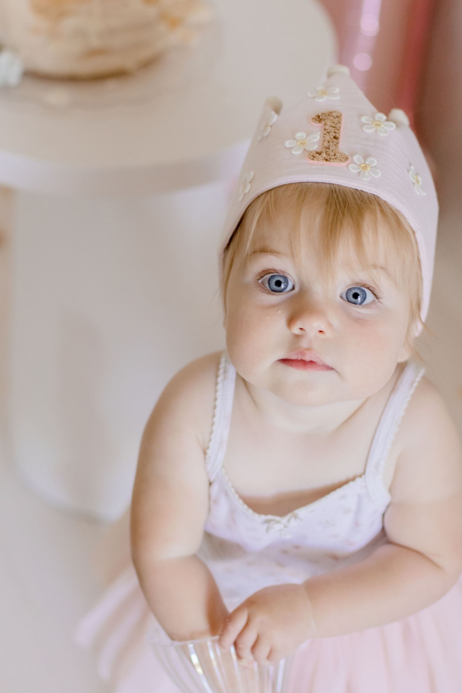 Baby in tulle dress and white crown with number 1 poses near white cake stand.