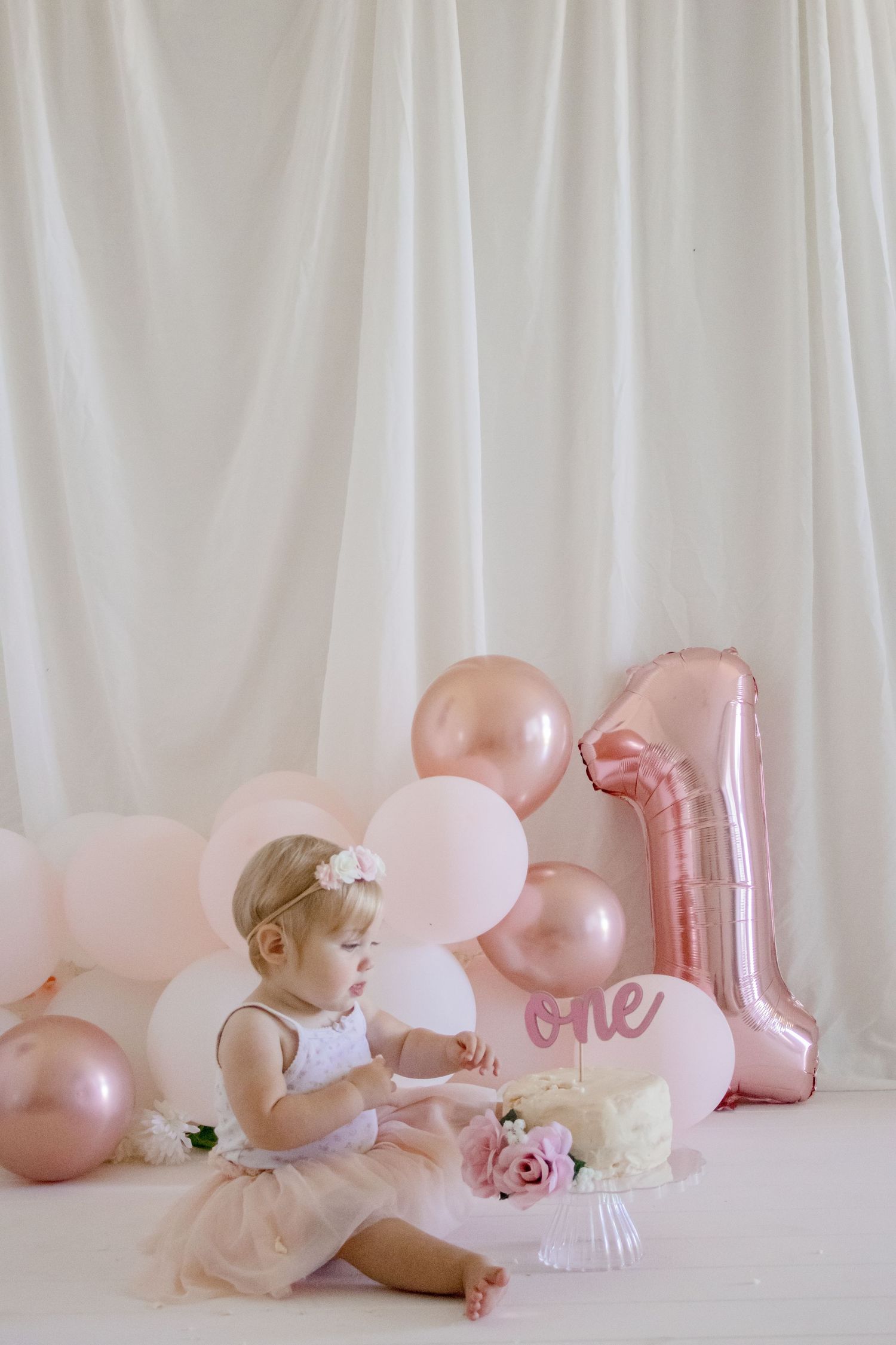 A first birthday celebration setup with pink balloons, roses and a white cake on a white backdrop.