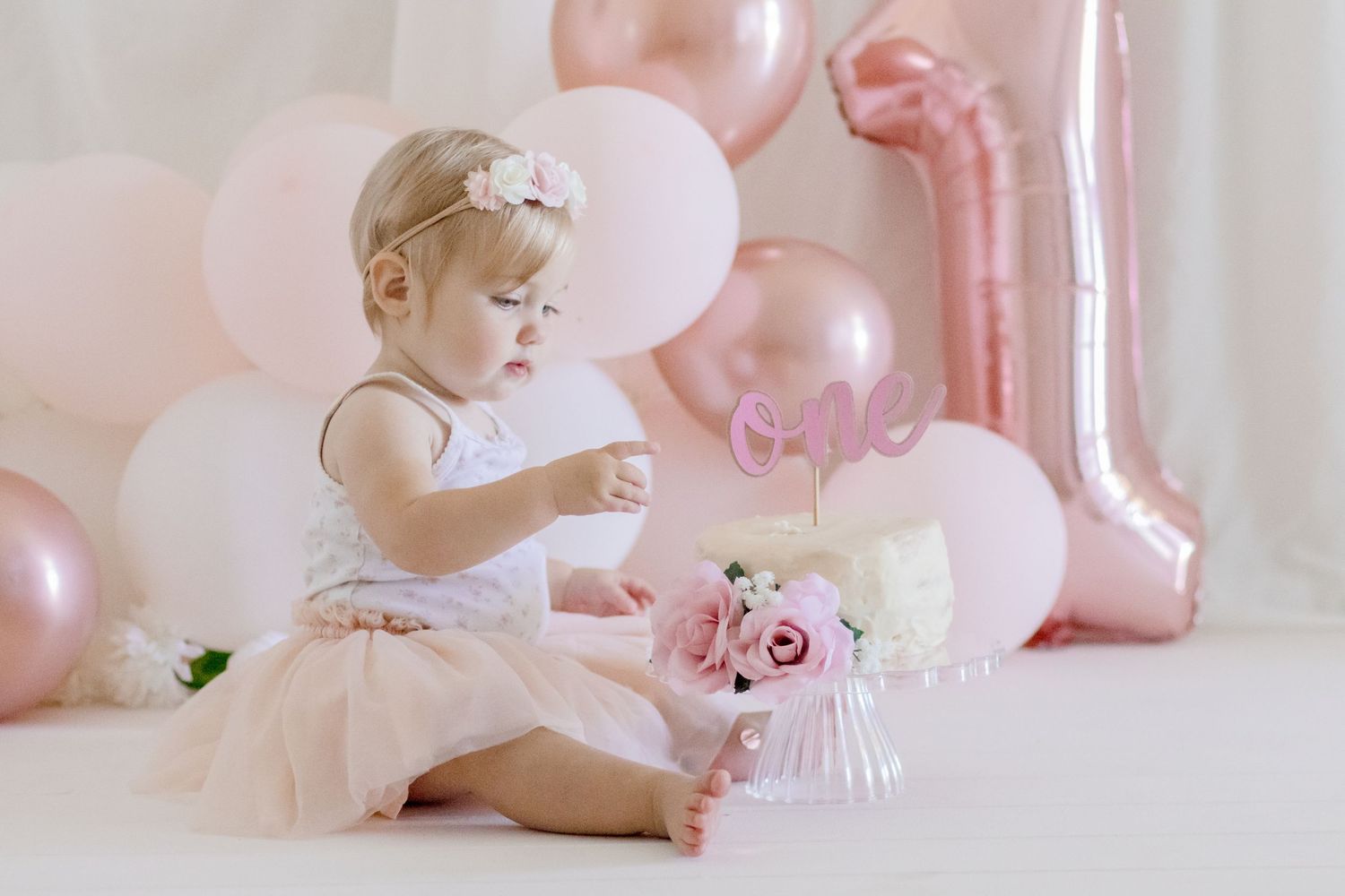 Baby in tulle dress sitting with birthday cake and pink balloons.