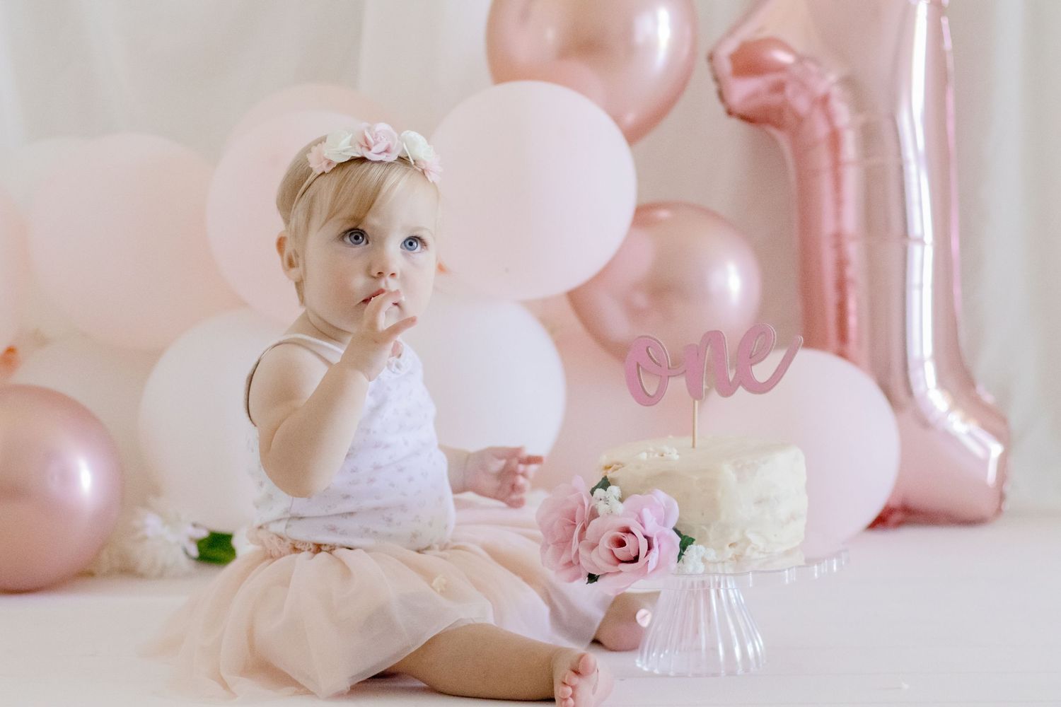 First birthday celebration setup with balloon decorations and white cake on pink background.