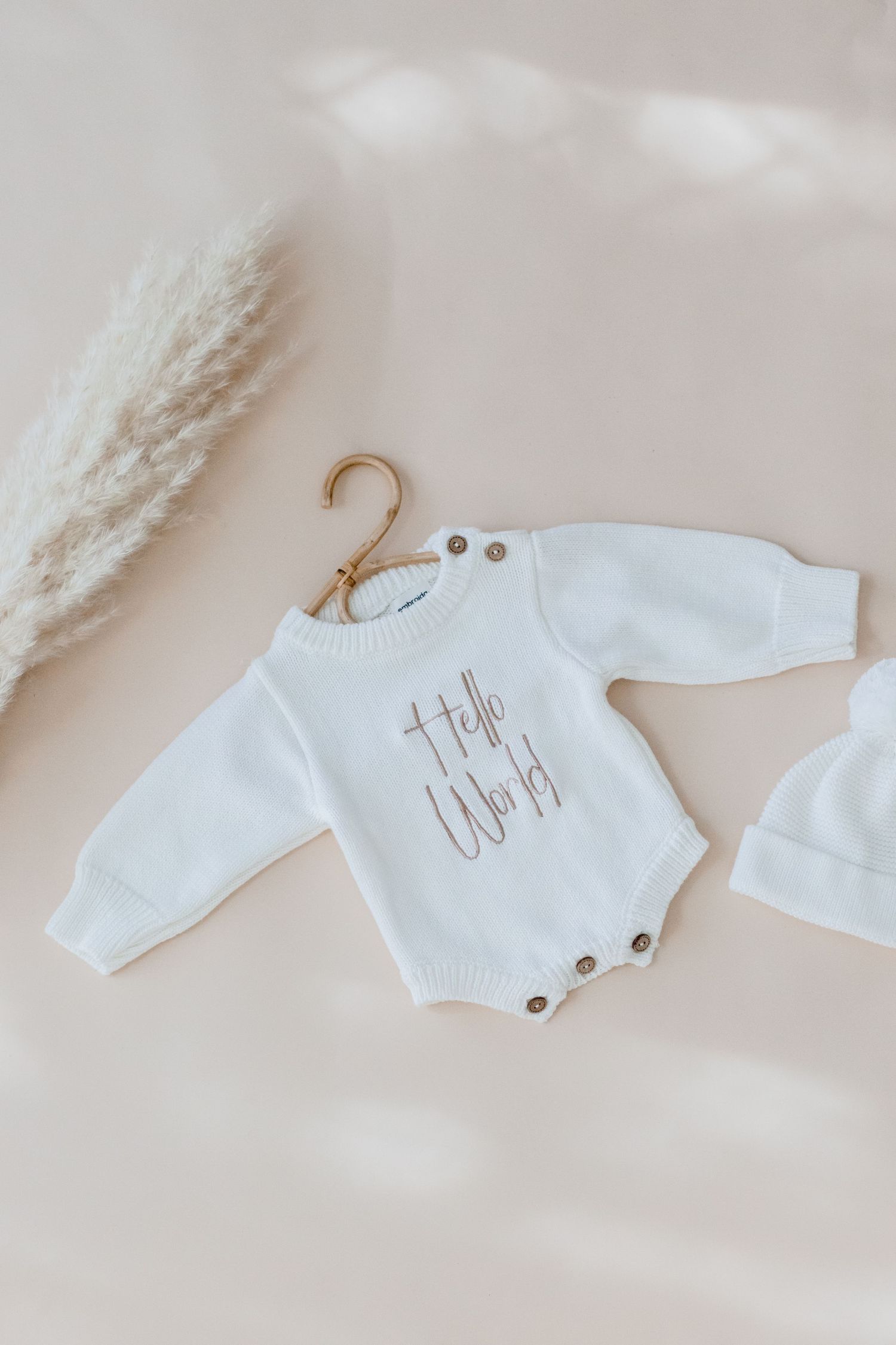White knit baby romper with text detail hanging on wooden hanger beside dried pampas grass on beige background.