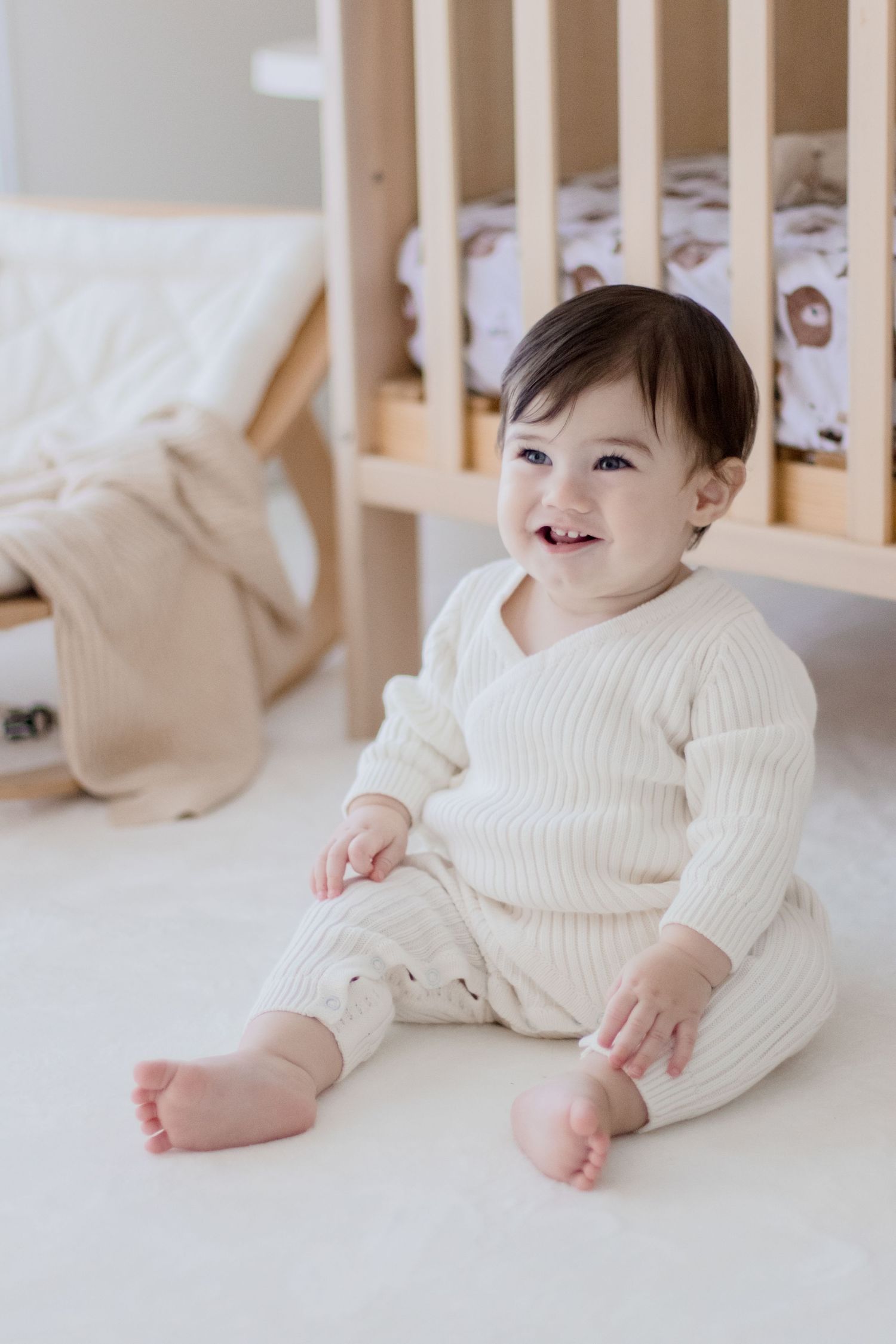 Baby in cream colored knit outfit sitting and smiling in wooden crib.