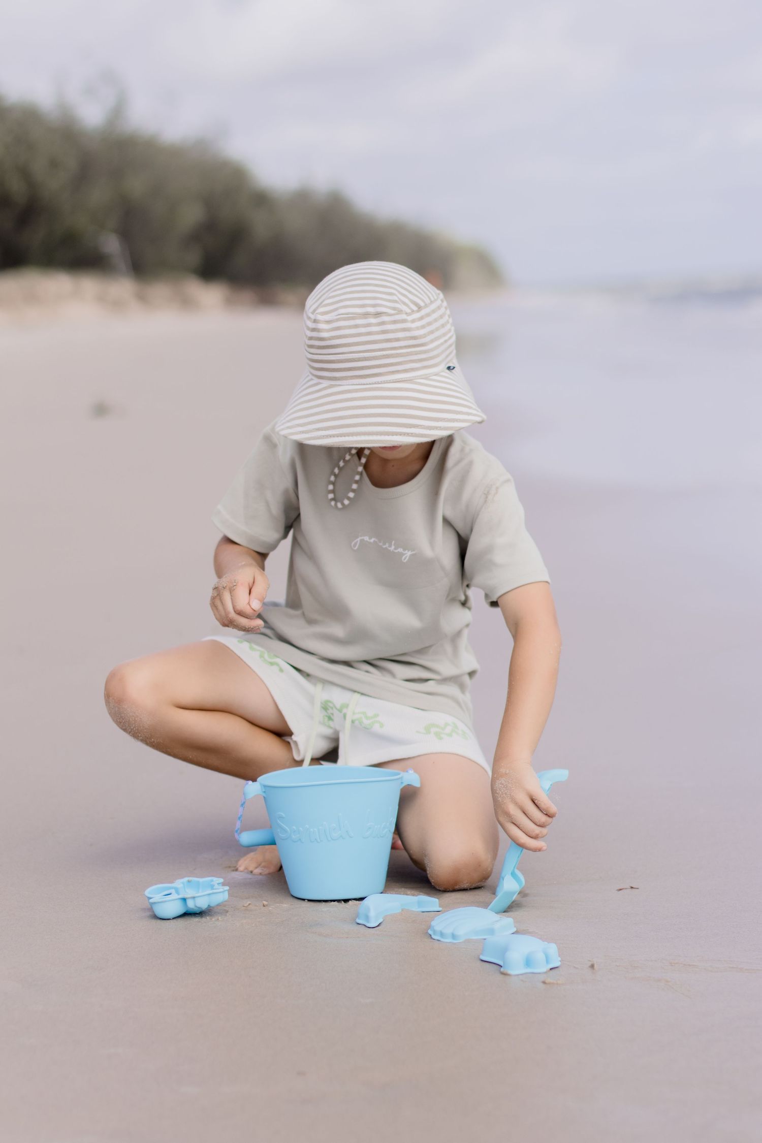 Young child playing with blue beach toys while wearing a sun hat on a sandy shoreline.