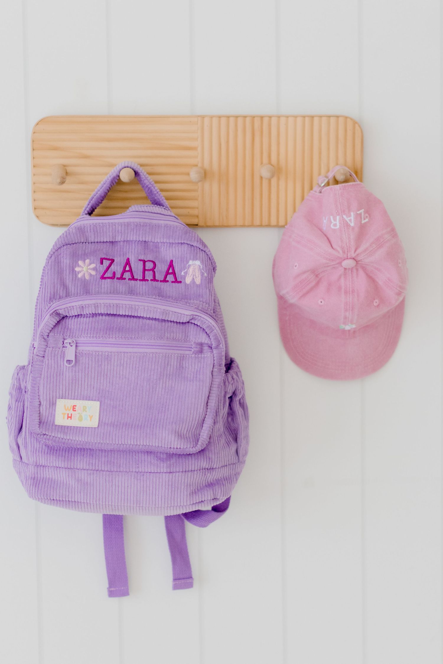 Purple Zara backpack and pink baseball cap hanging on wooden wall hooks.