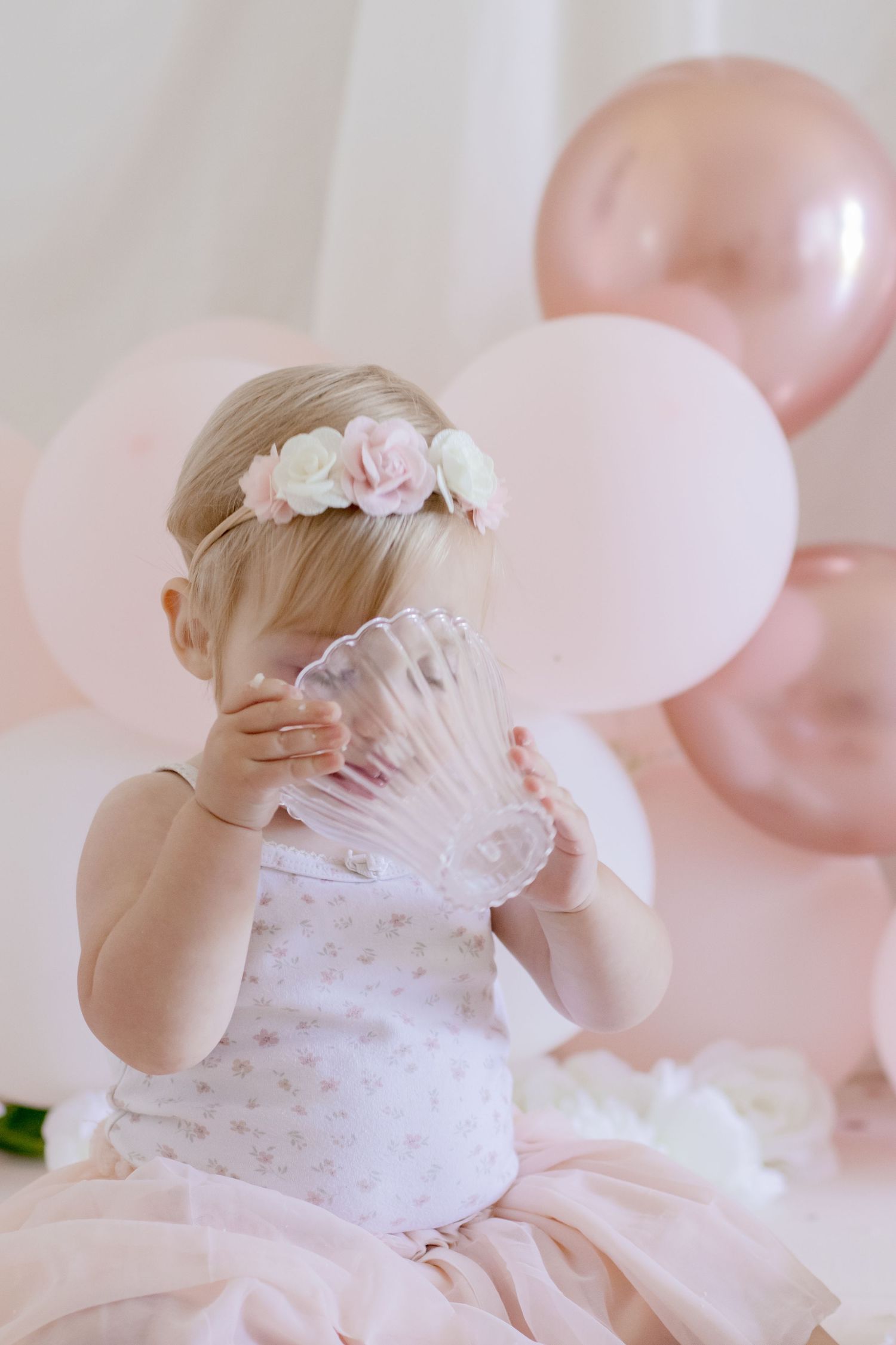 Baby in tulle dress and floral crown holding crystal glass with pink balloons in background.