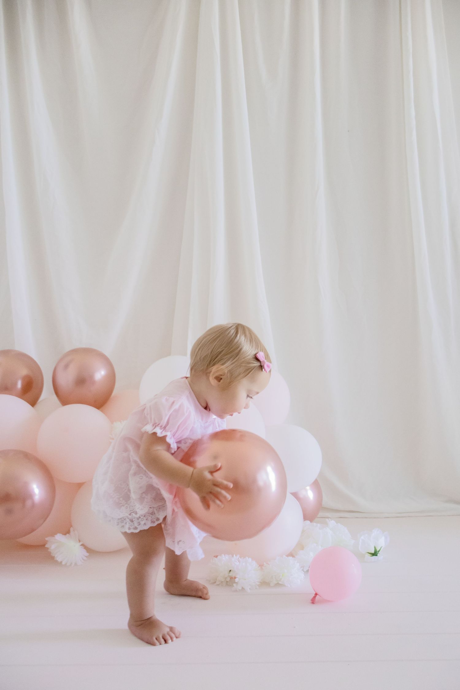Small child in pink dress bending down to touch rose gold balloons on white floor.