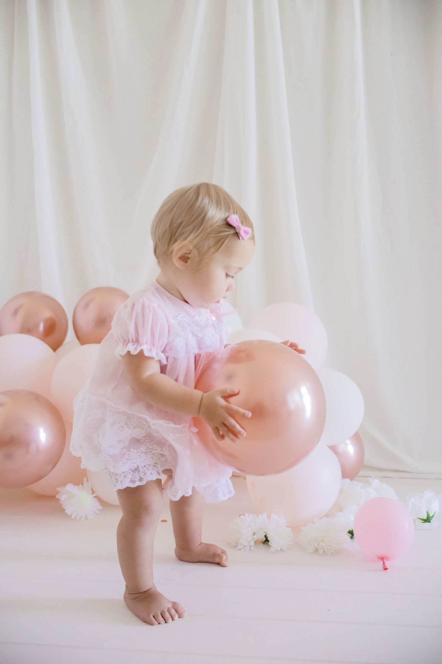 Baby standing in pink dress reaching for metallic pink balloons with white curtain backdrop.