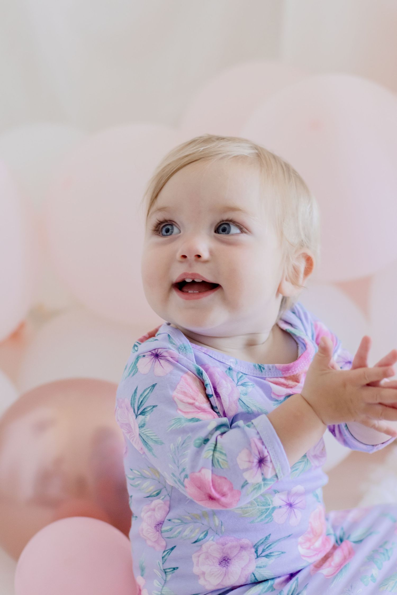 Smiling baby in floral outfit looks up while surrounded by pink balloons.