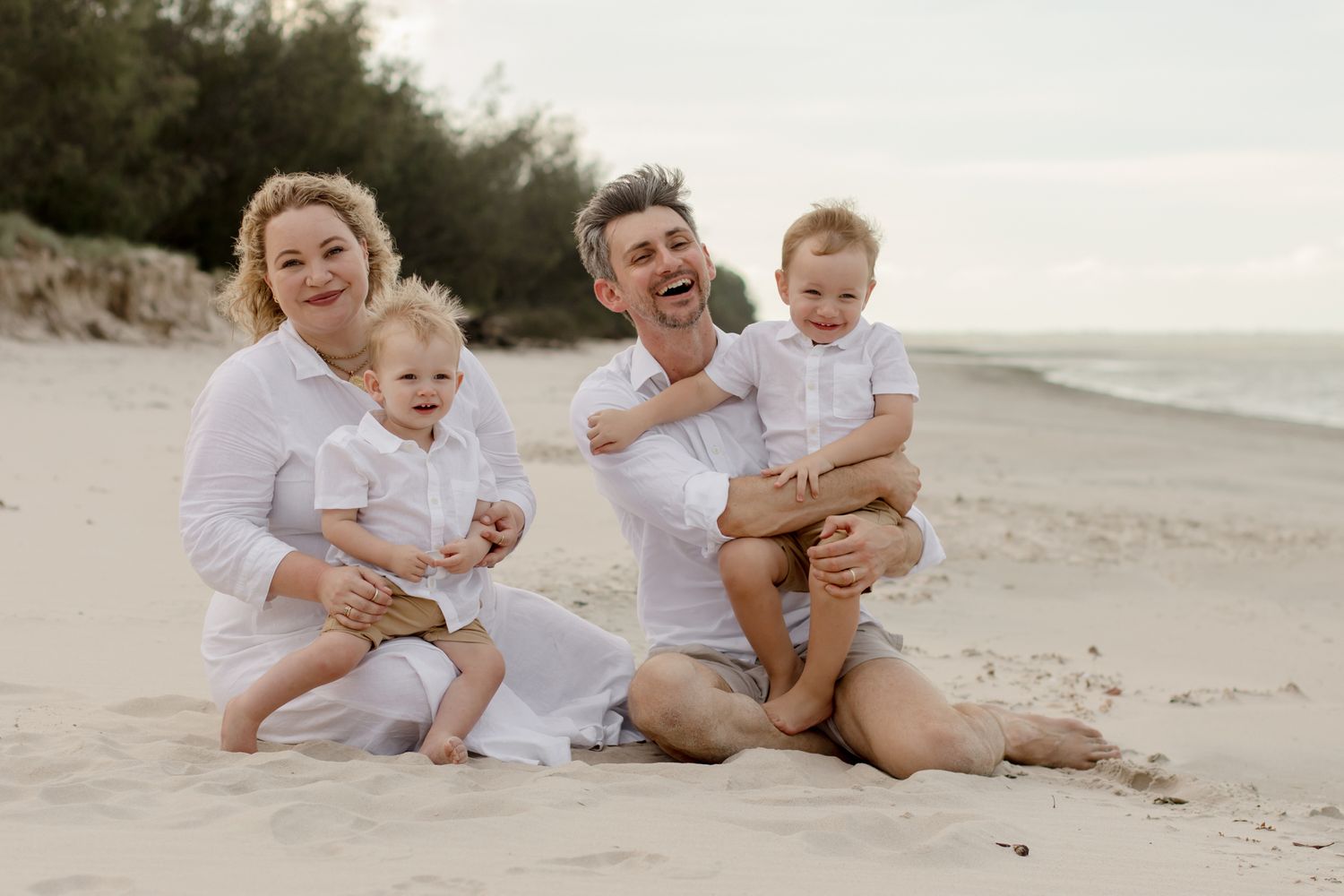 A cheerful family wearing white sits together on a sandy beach.