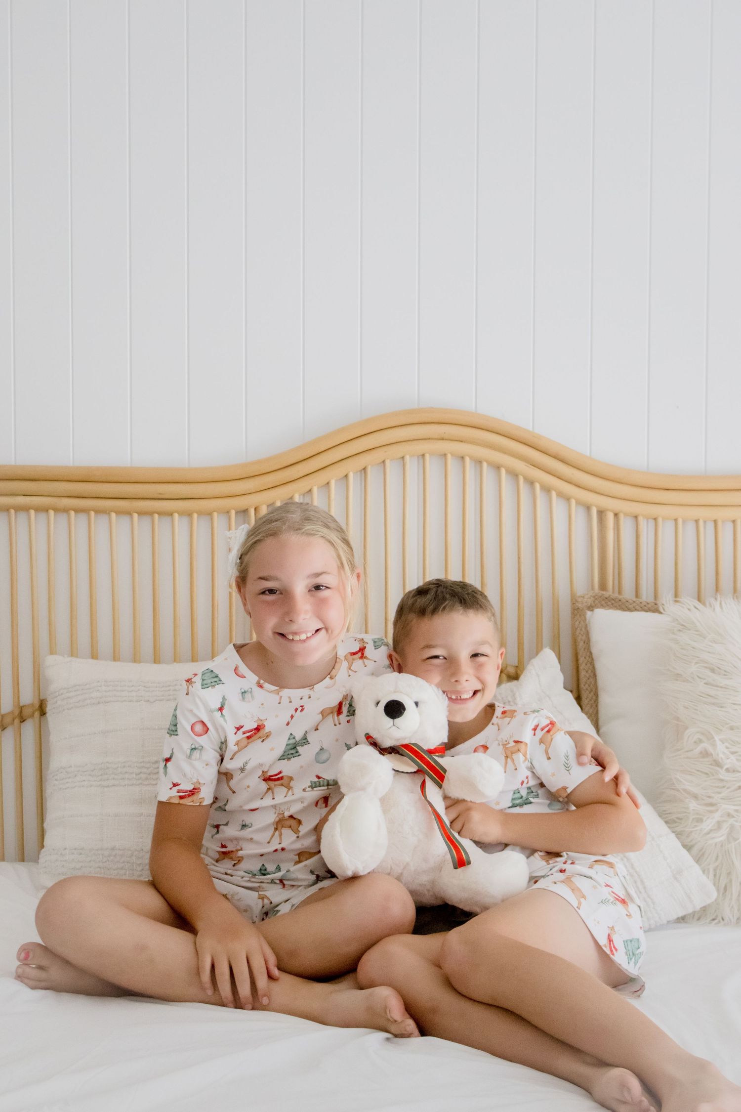 Children in matching floral pajamas cuddle with white teddy bears on a wooden bed with white bedding.