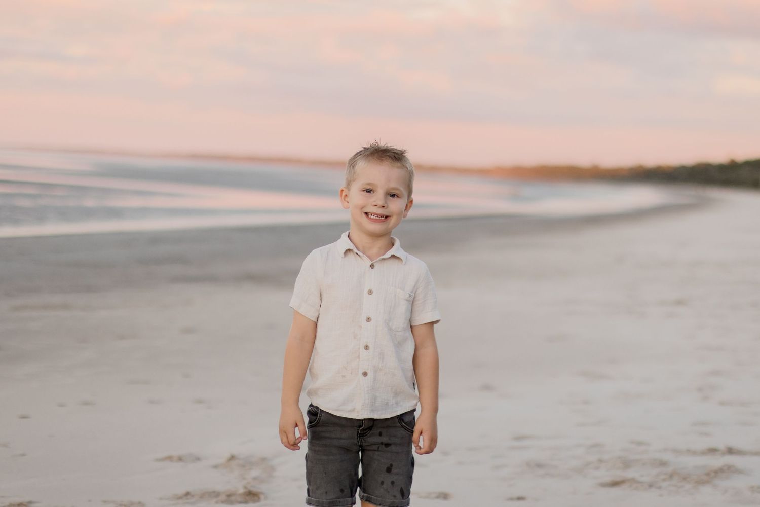 A figure in a white button-down shirt and dark pants stands on a peaceful beach at sunset.