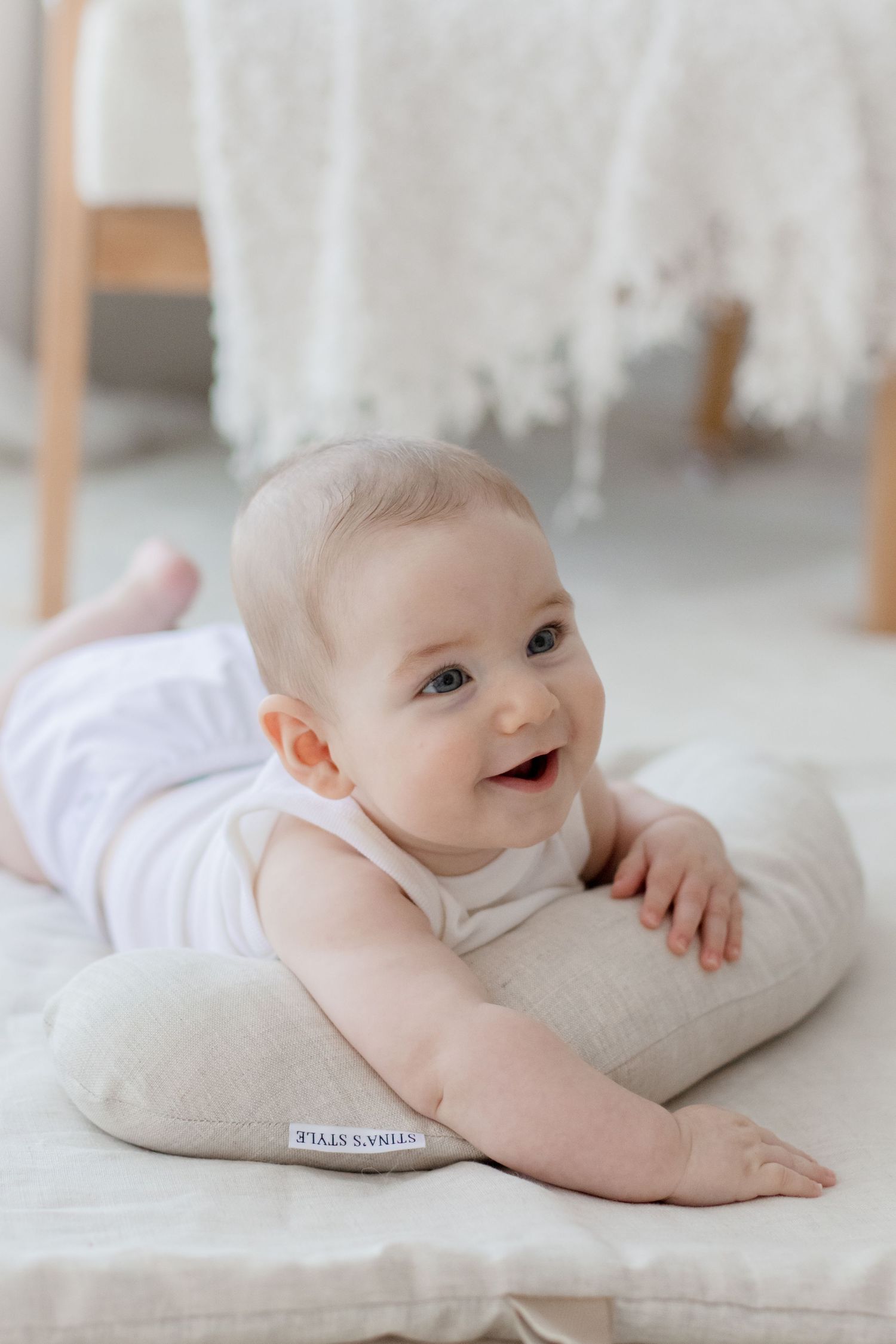 A happy baby in white clothing lays on a beige cushion, smiling at the camera in a bright room.