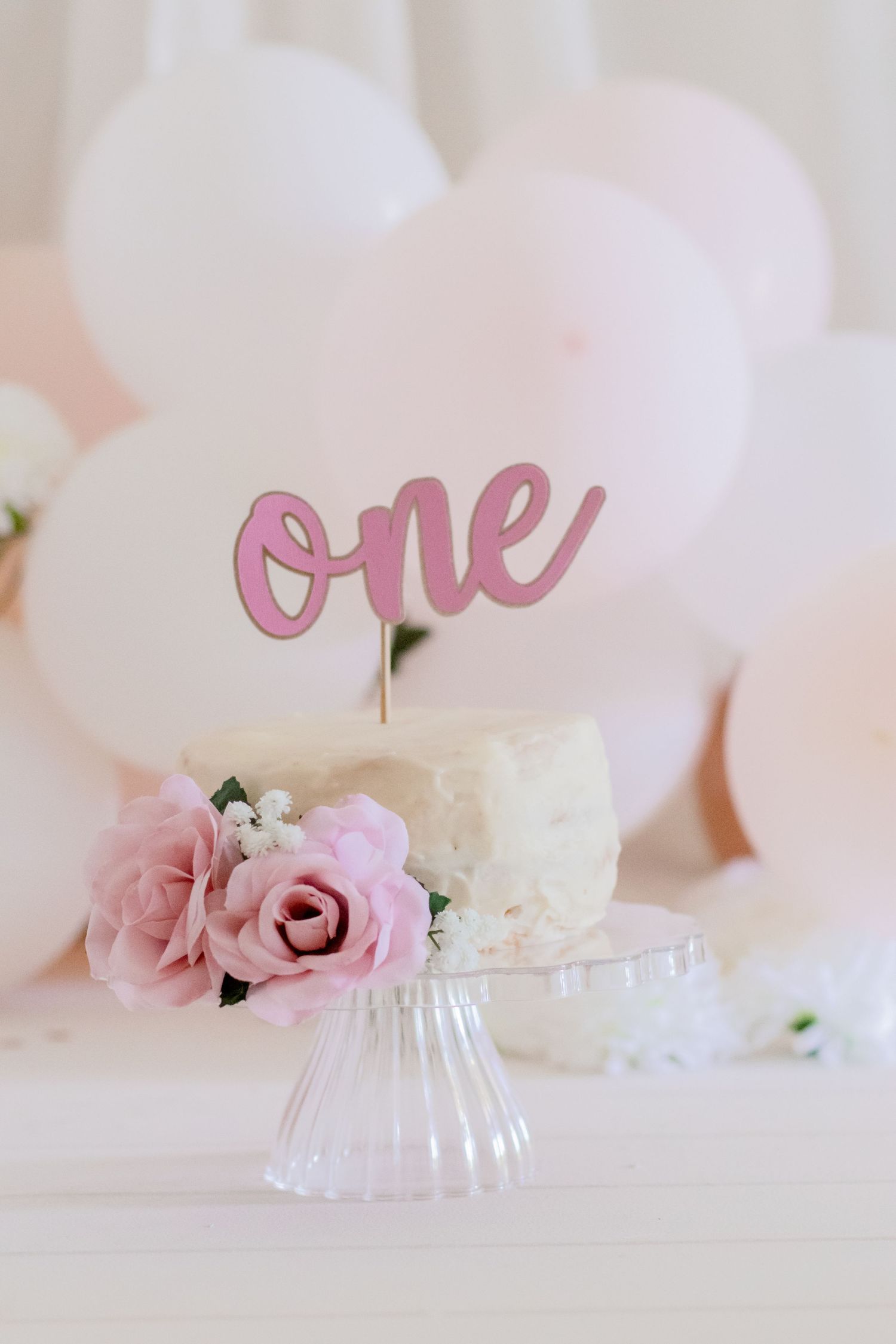 White birthday cake decorated with pink roses and 'one' cake topper on glass stand.