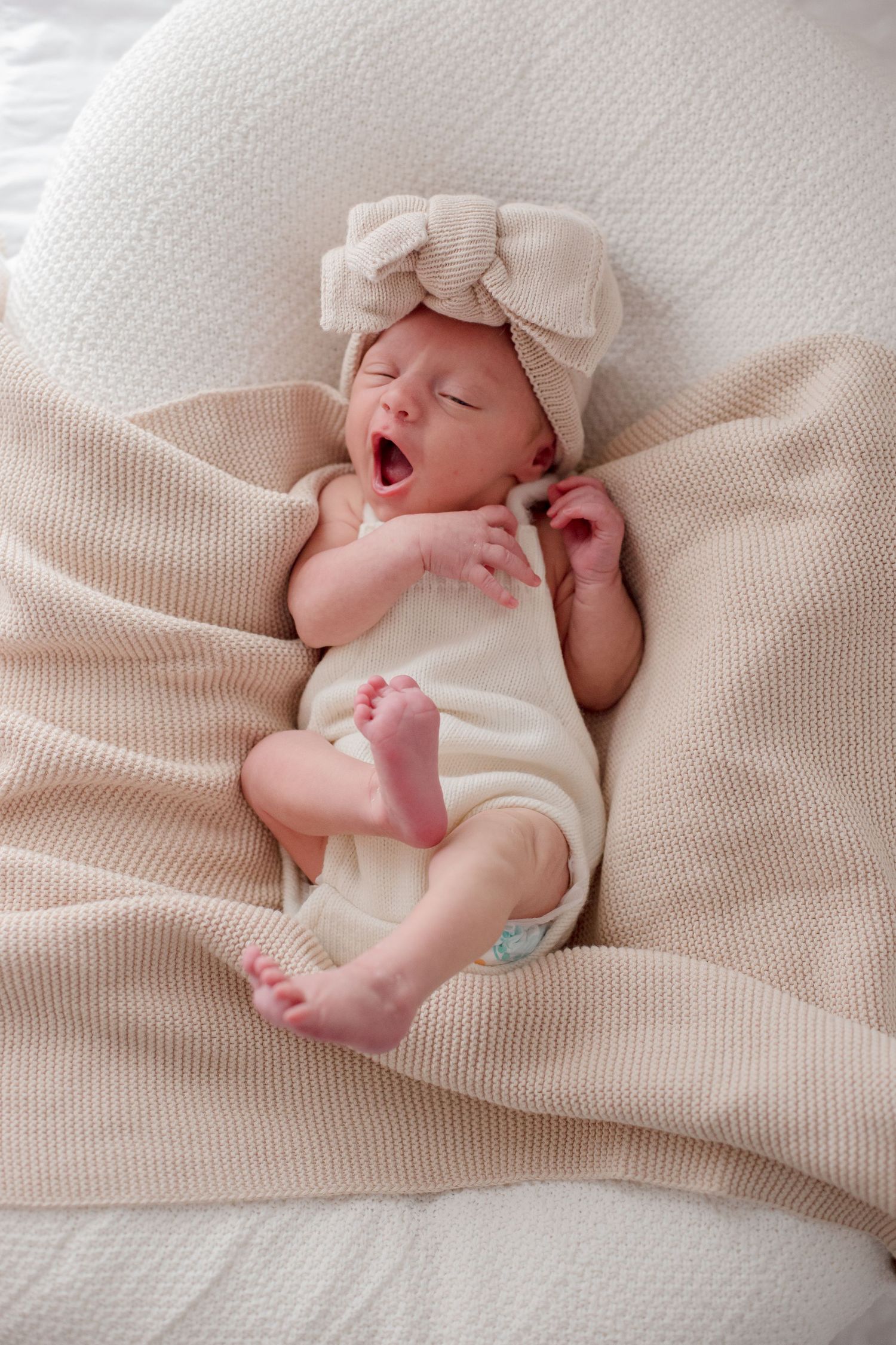 Newborn baby in cream outfit and bow headband yawns while lying on textured blanket.