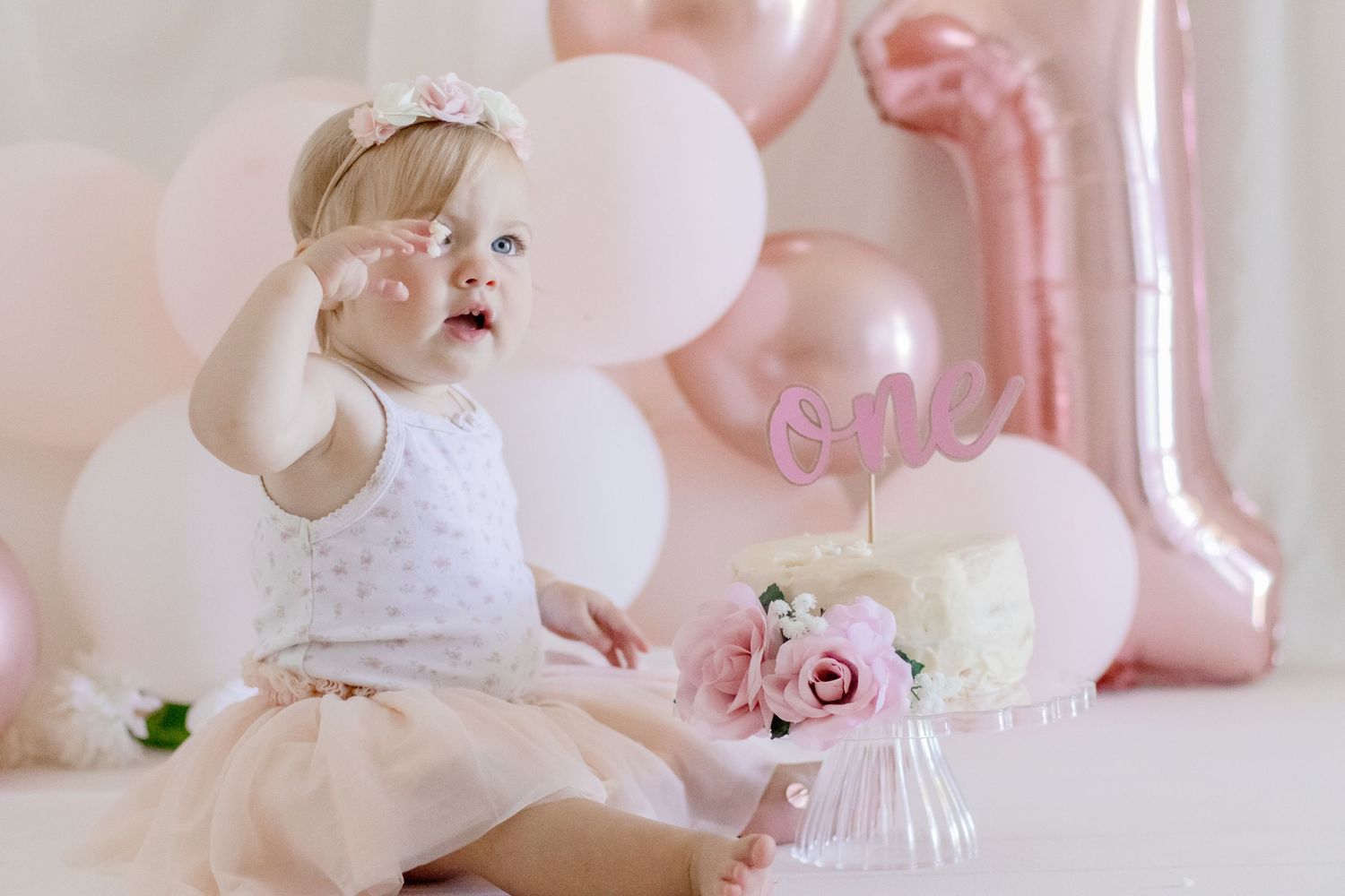 Baby gesturing during first birthday celebration with pink balloon decorations.