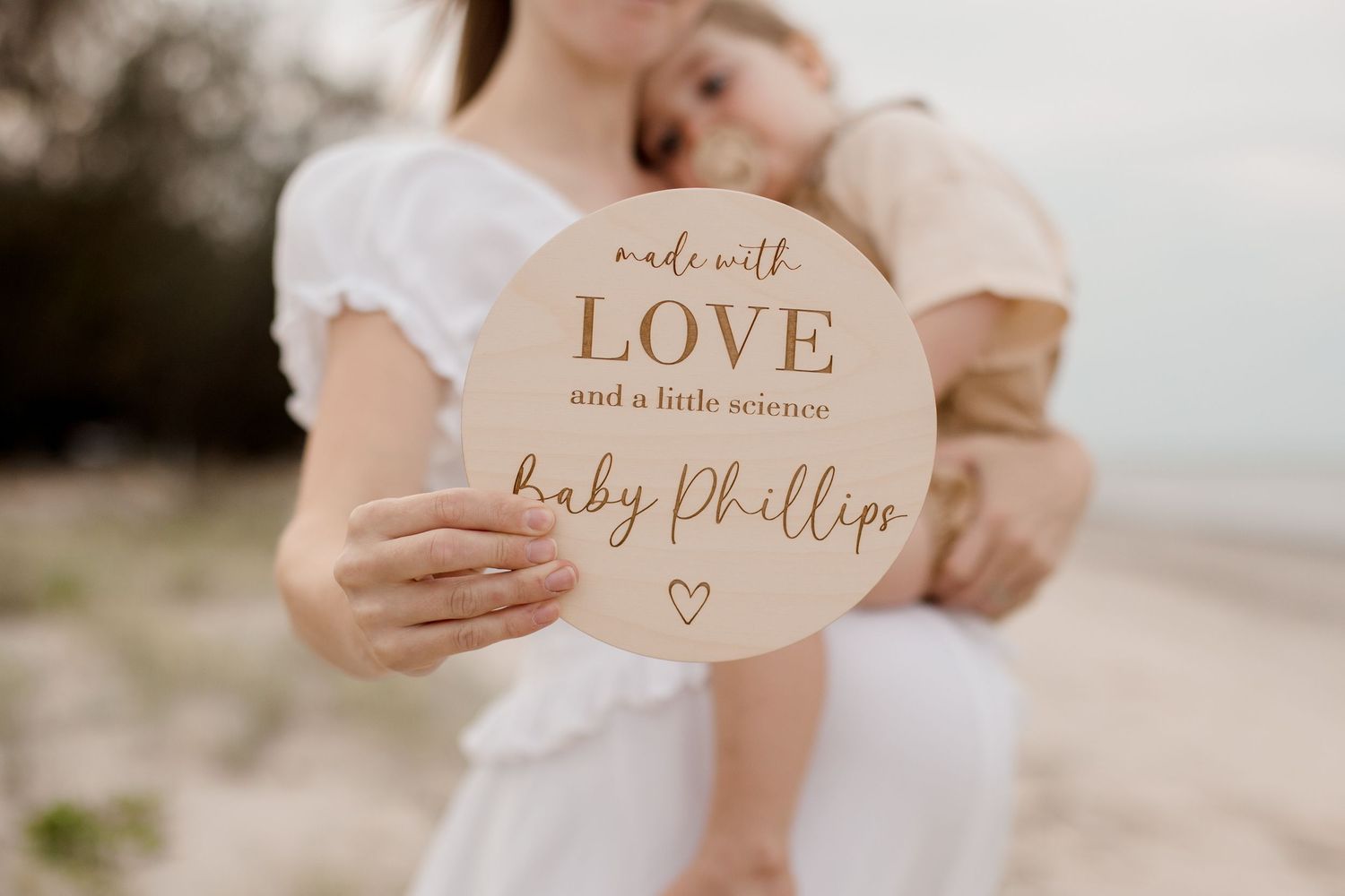 Hand holding circular wooden baby announcement sign outdoors.