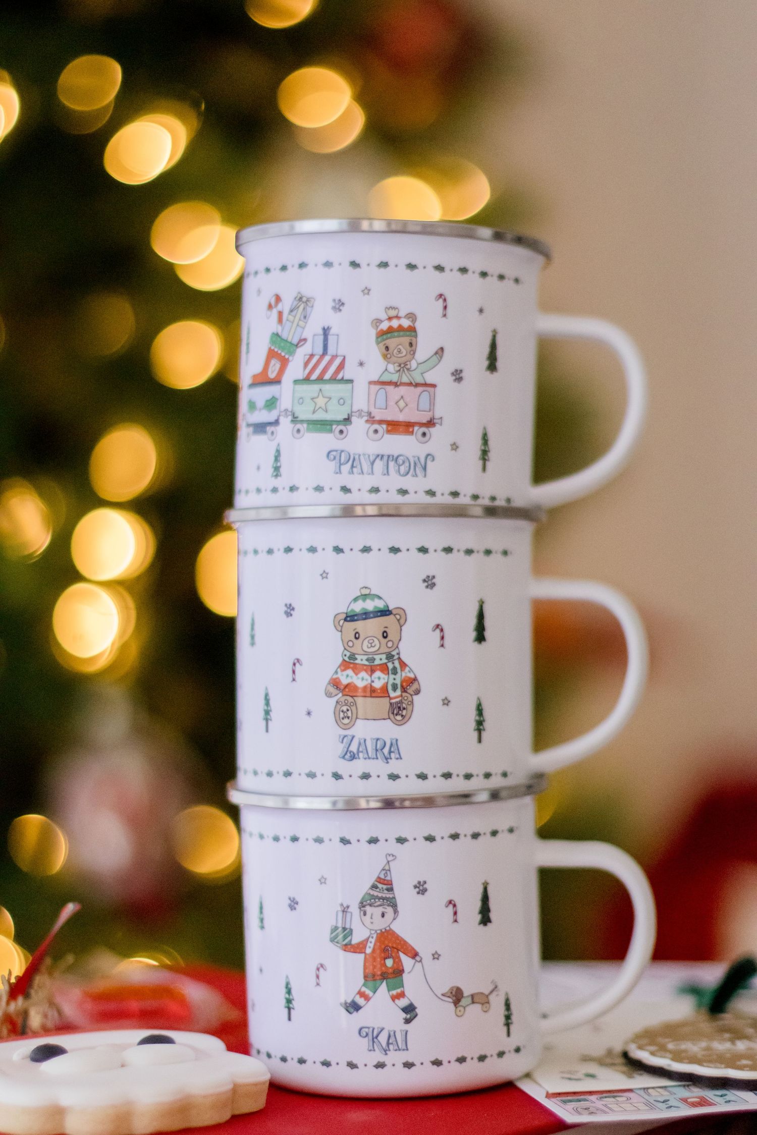 White ceramic mugs with whimsical Christmas village illustrations stacked against a bokeh light background.