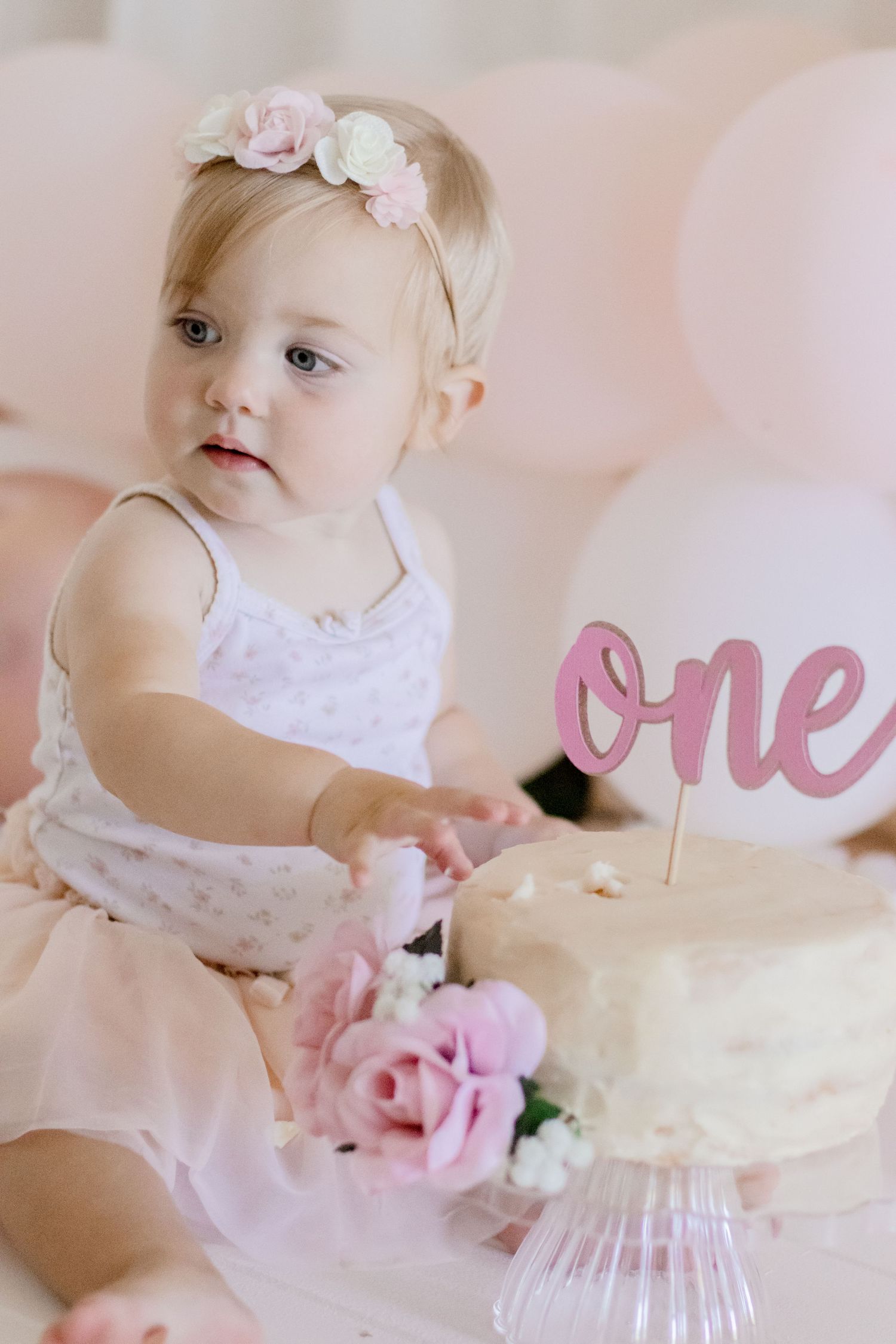 Baby wearing floral headband next to birthday cake with pink 'one' cake topper.