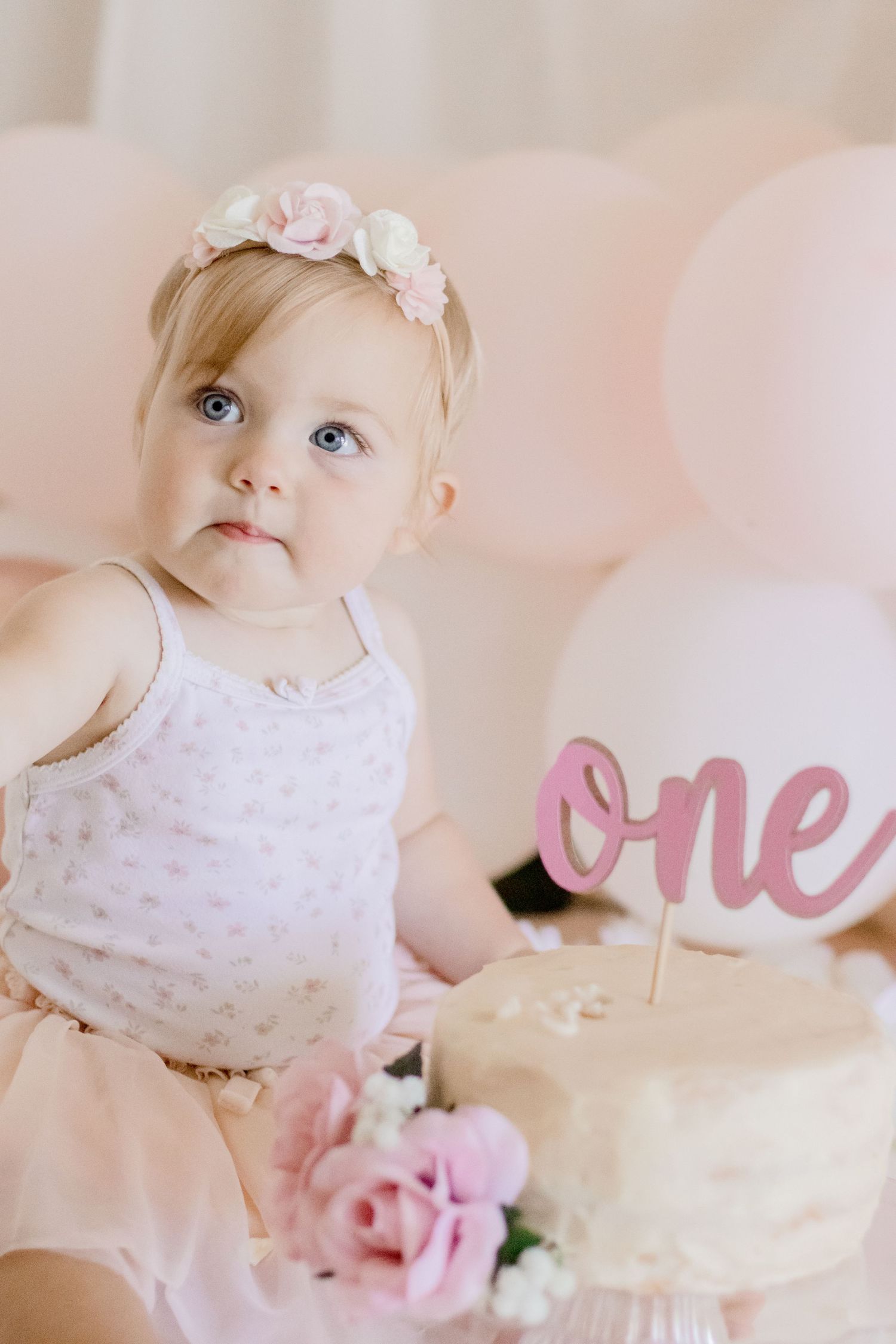 Baby in tulle outfit looking away from camera next to birthday cake with pink decorations.