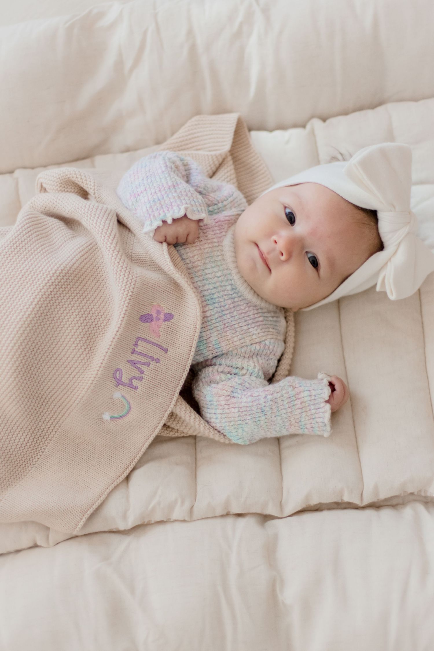 Baby lying on beige bedding wearing a soft knit sweater in pastel colors.