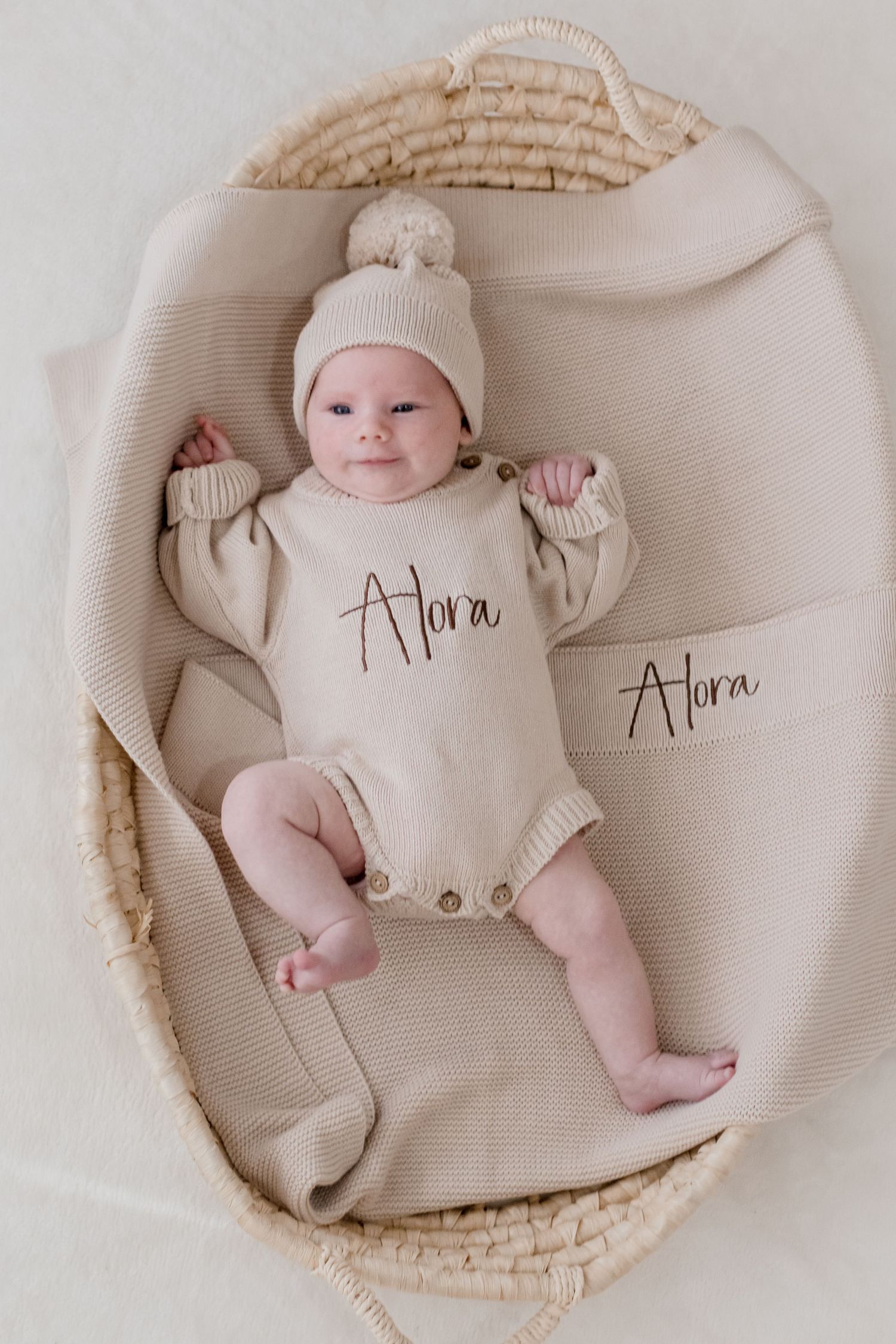 Adorable newborn baby wearing white outfit lying in woven basket with personalized 