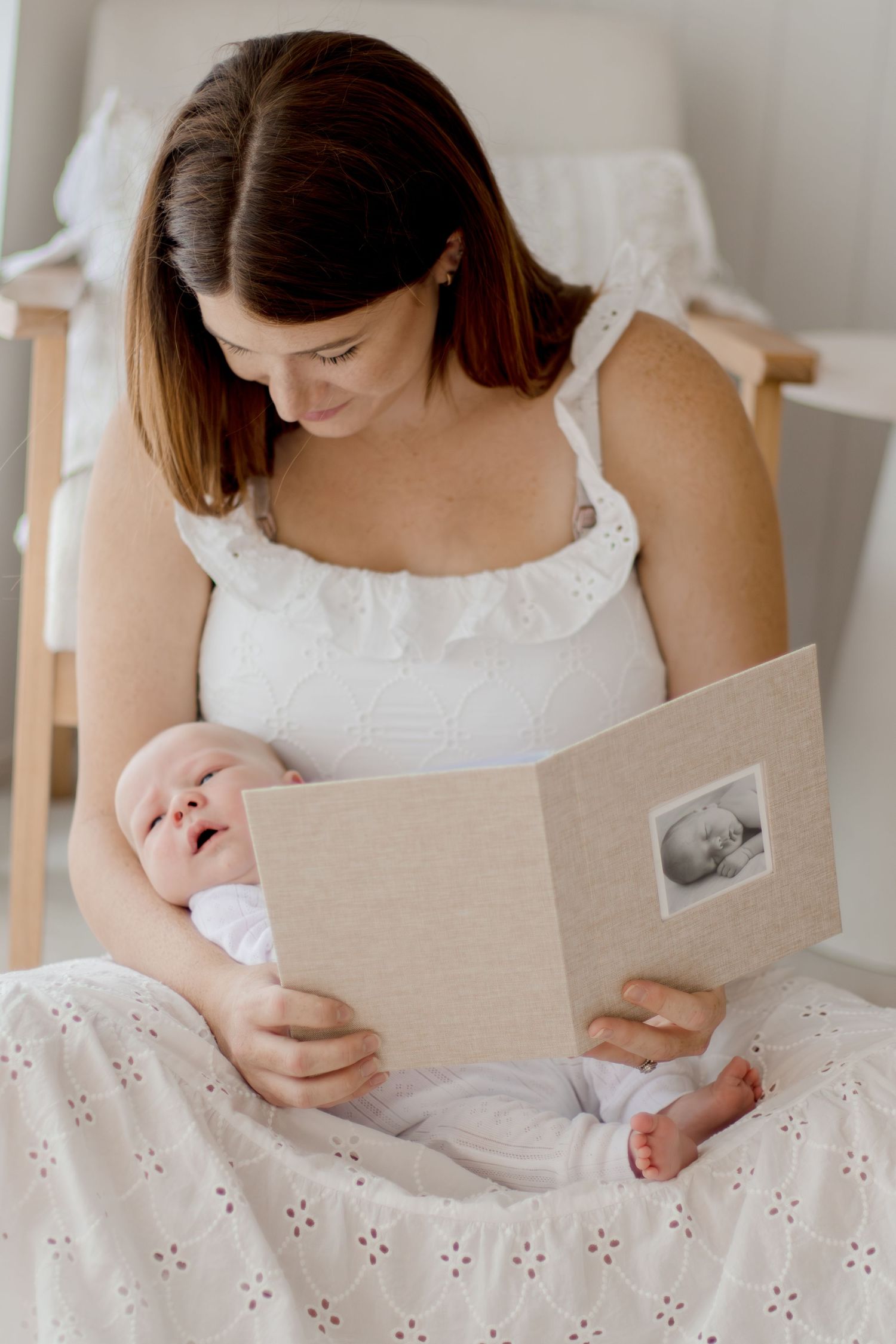 A person in a white eyelet dress reads from a book while sitting on a bed with white linens.