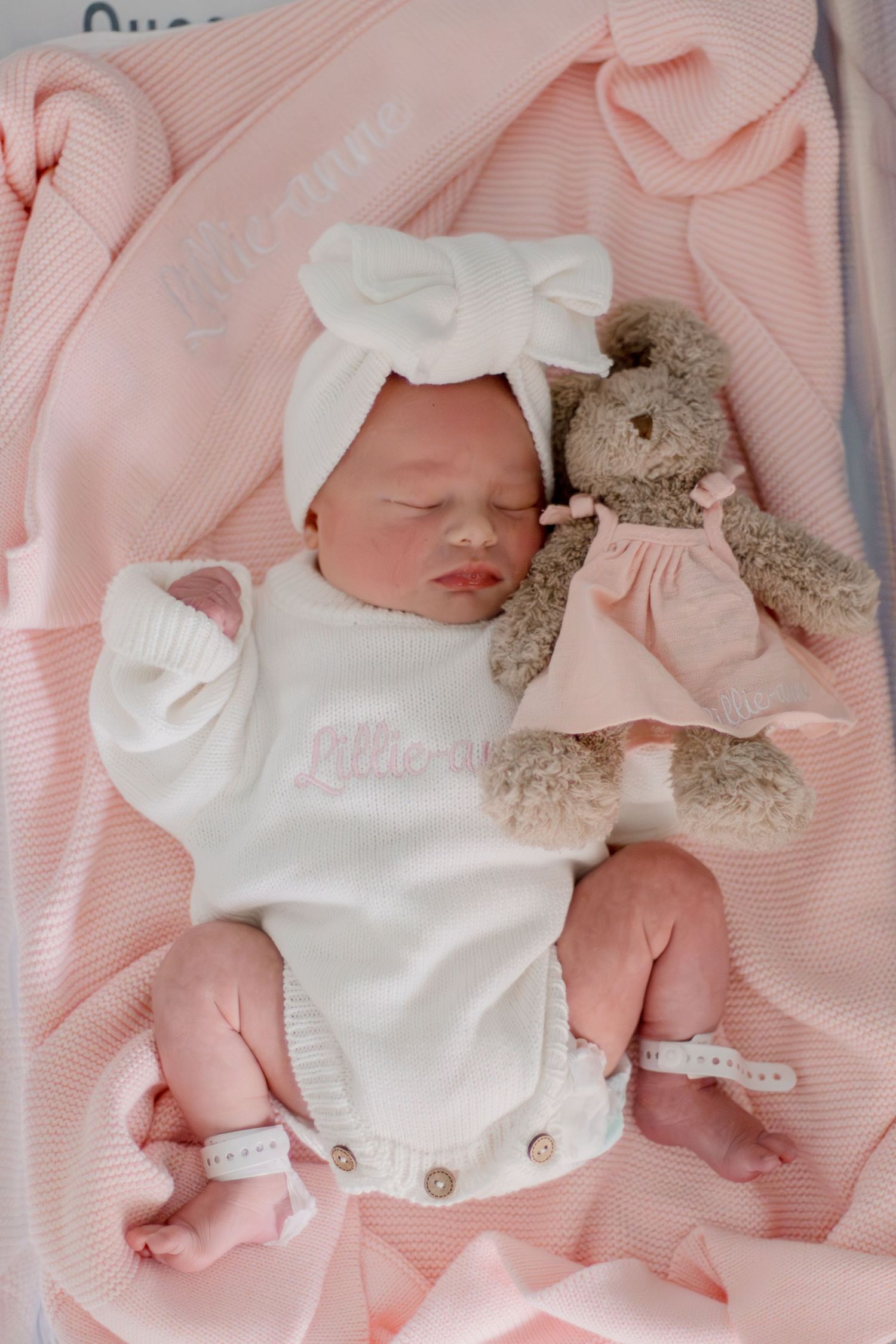 Sleeping newborn baby in white onesie with bow headband beside teddy bear on pink blanket.