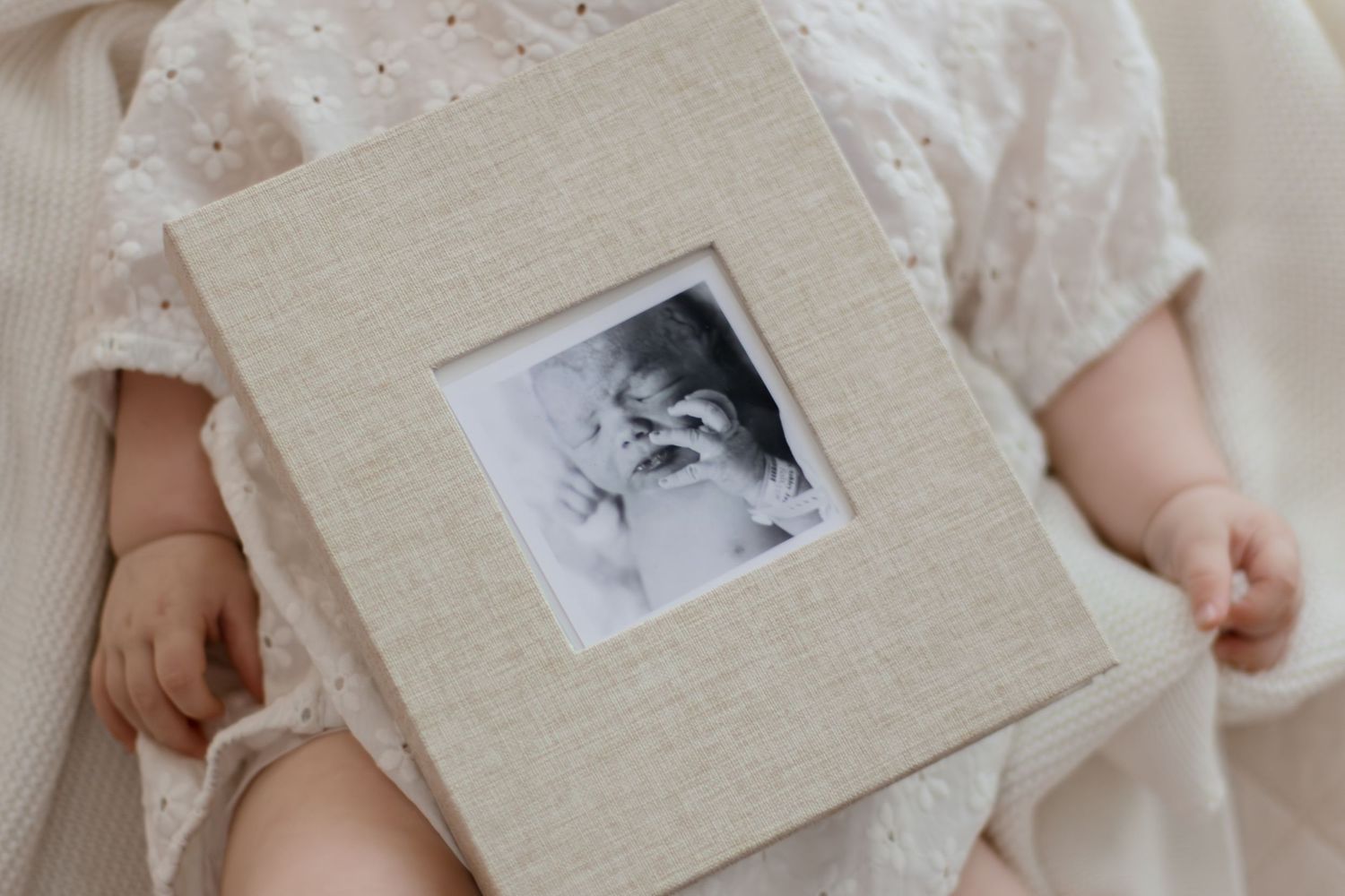 Baby in white dress holding a linen photo album with black and white infant portrait on cover.