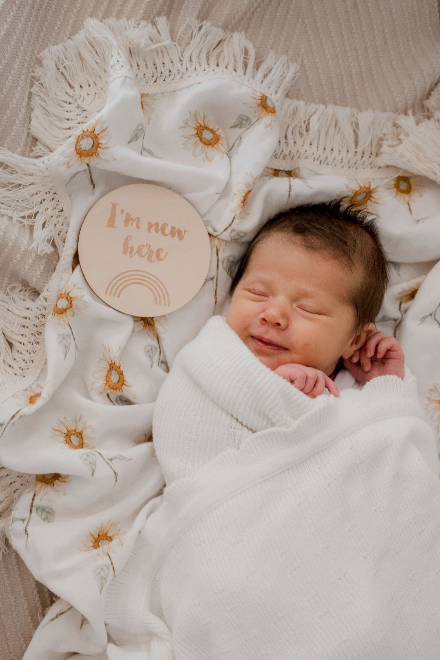 Sleeping newborn wrapped in white blanket on floral bedding with wooden 'I'm new here' sign decoration.