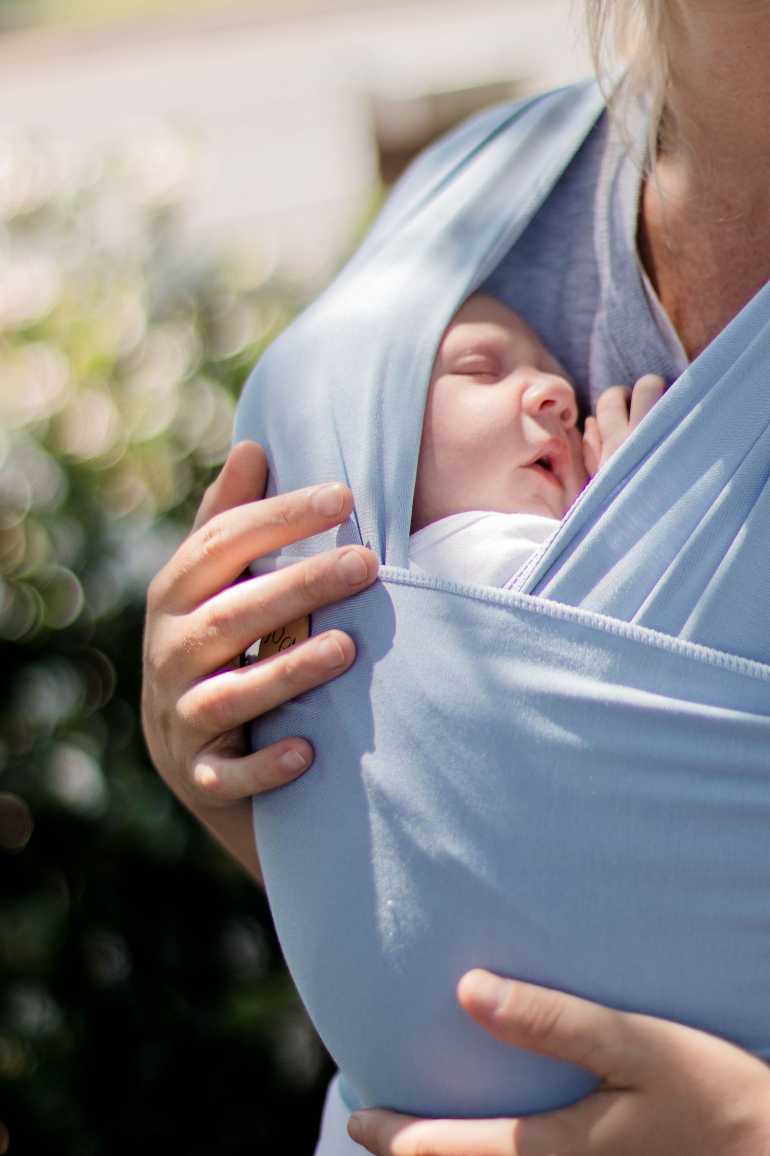 A series of images showing how to adjust and position a light blue baby carrier sling against a blurred background.