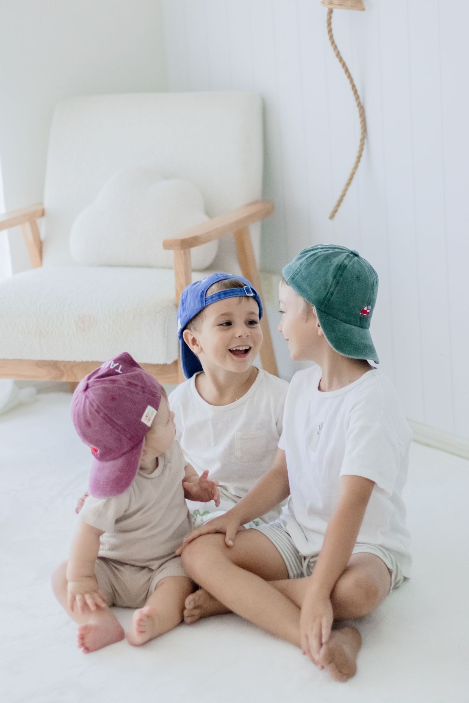 Three children wearing baseball caps sitting together and laughing on white floor.