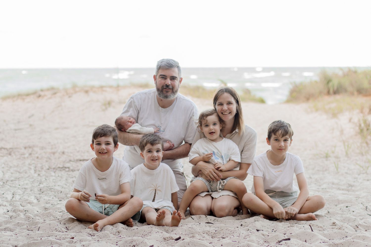 Family of six sitting together on a sandy beach in coordinated white and neutral outfits.