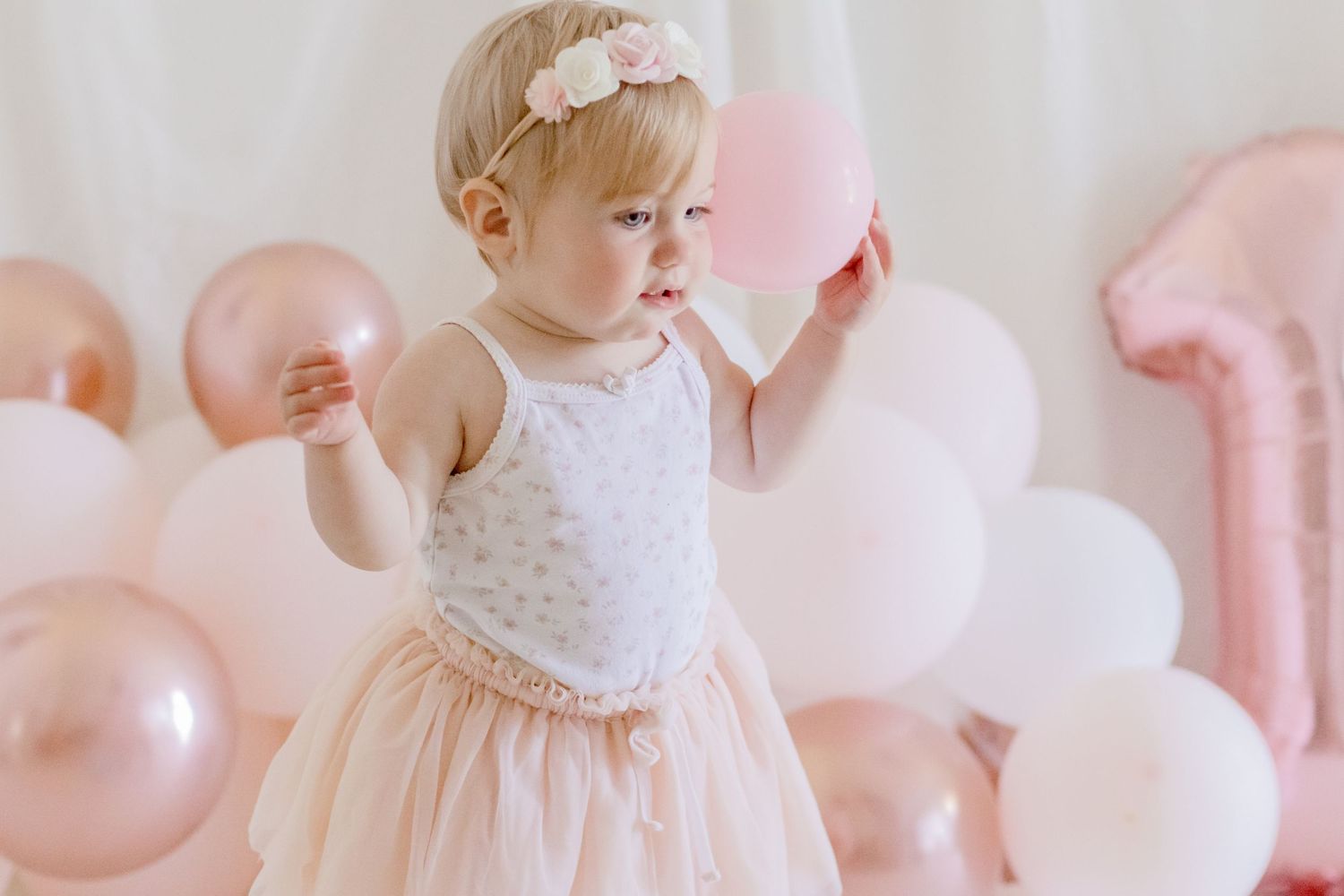 Baby in white dress posing with pink and rose gold balloons for first birthday photos.