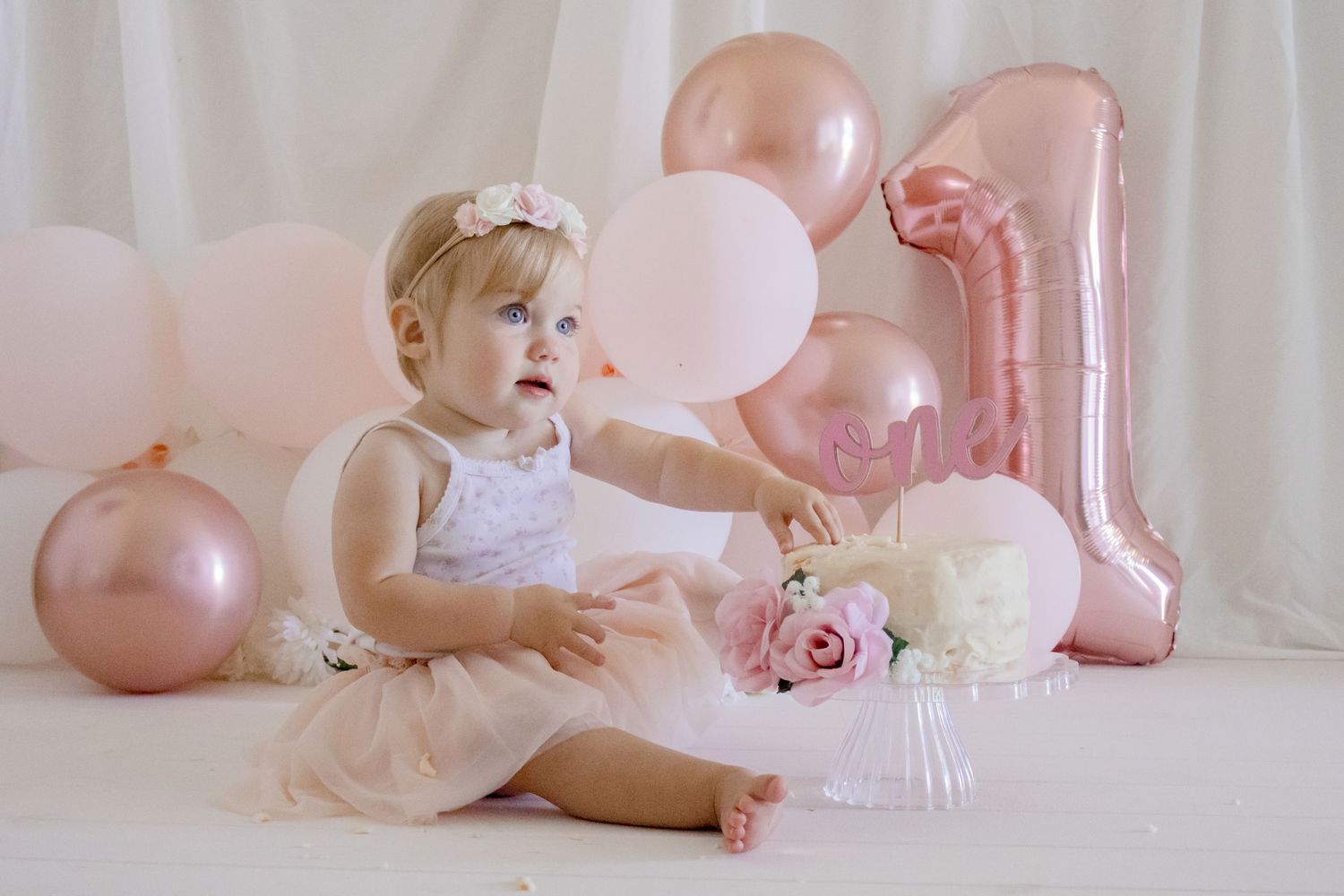 A first birthday celebration with pink balloons, roses, and white cake against a white curtain backdrop.