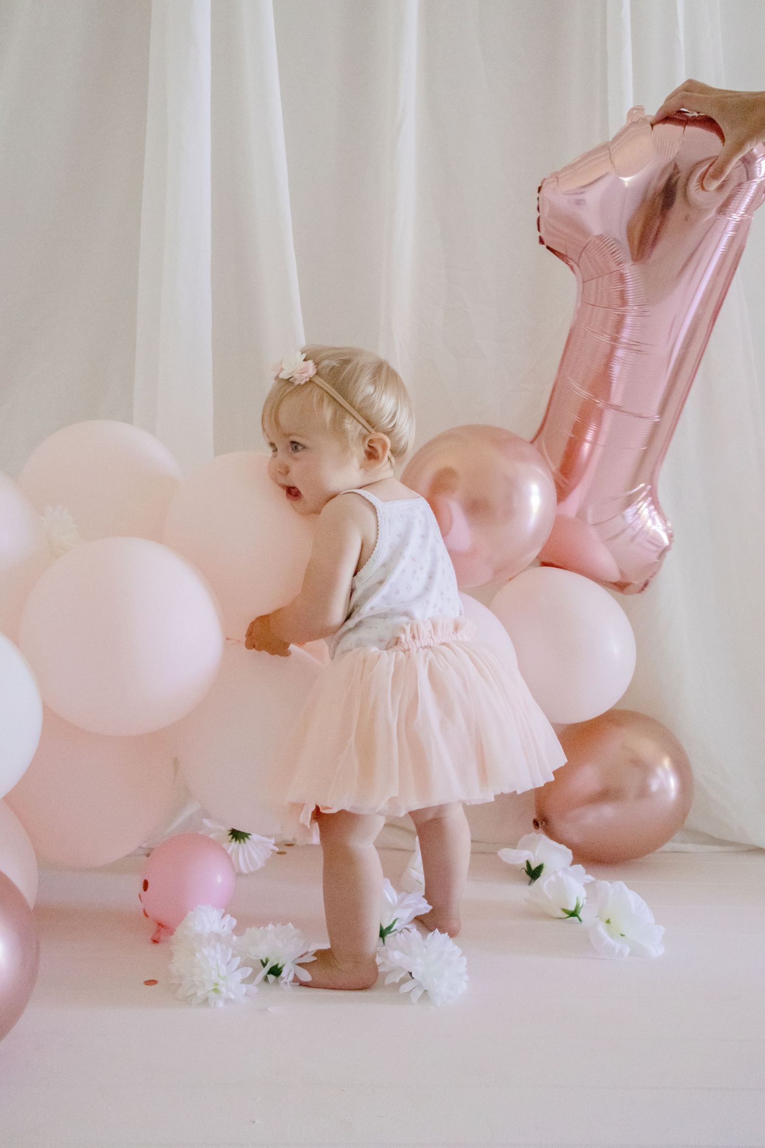 A toddler in a tulle dress explores pink and rose gold party decorations against a white backdrop.