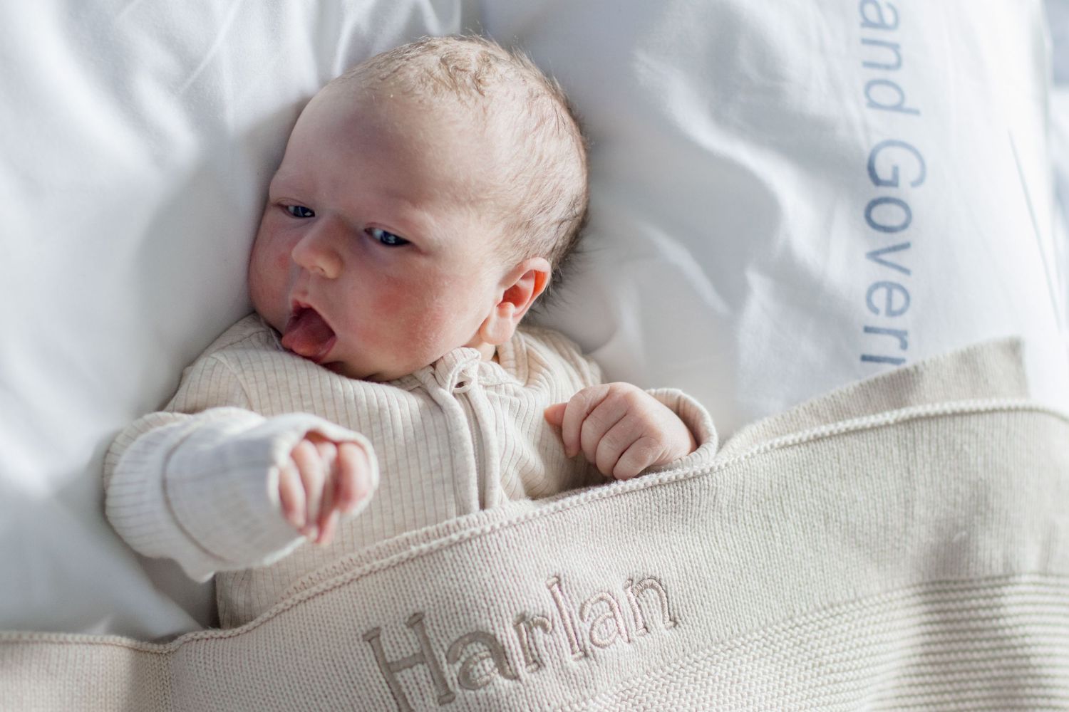 A newborn baby in a white knit outfit yawning while lying on monogrammed bedding.