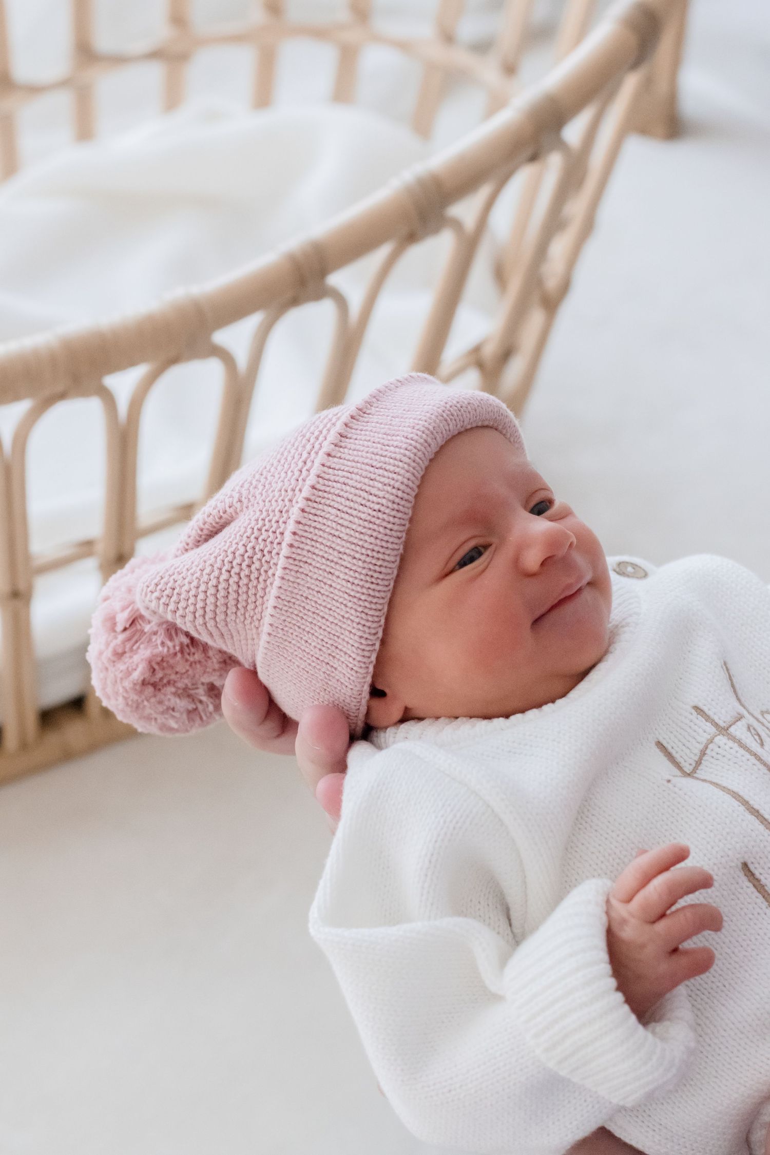 Newborn baby wearing soft pink knit hat lying on white bedding with rattan chair in background.