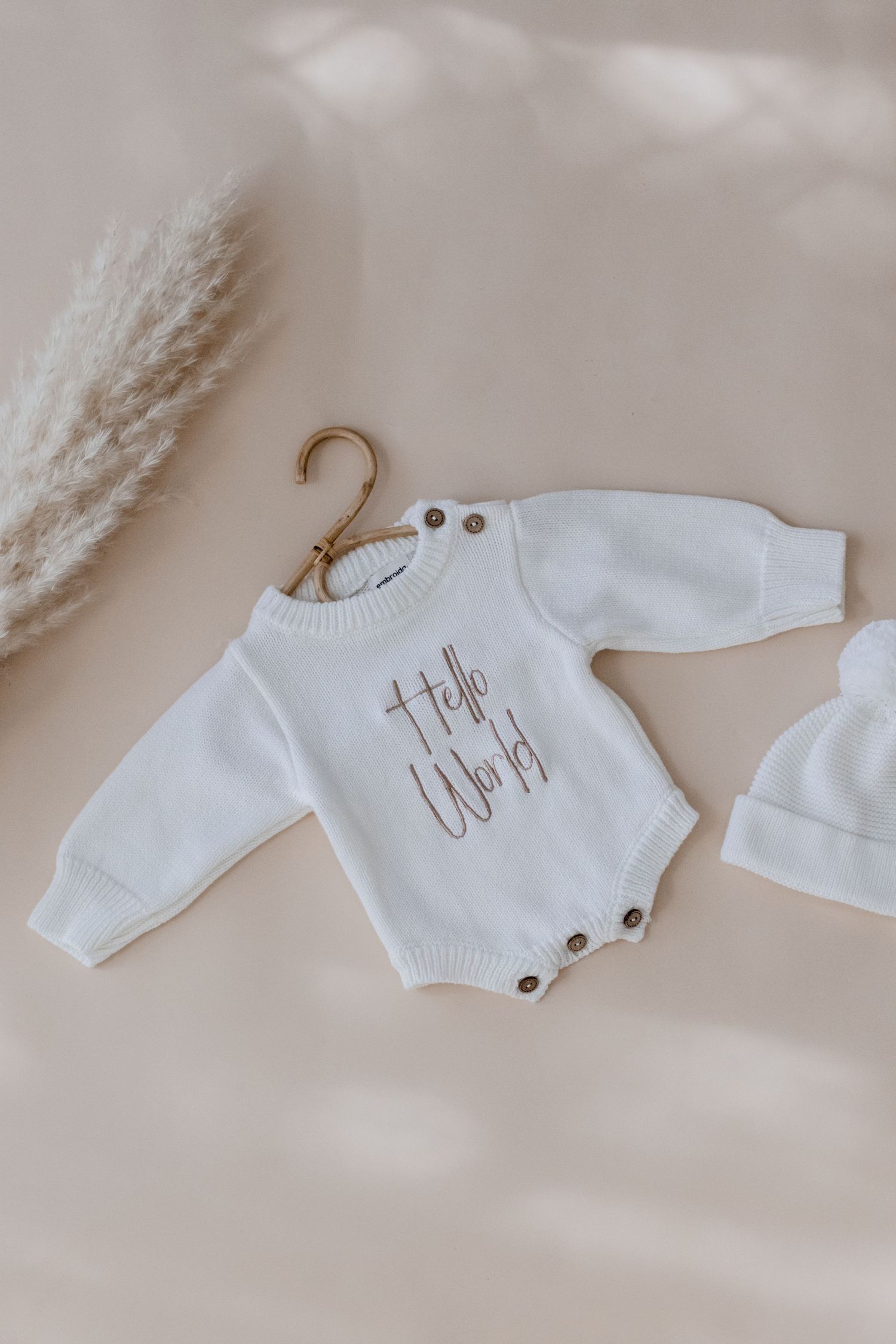 White knit baby romper with text design displayed on wooden hanger next to decorative dried pampas grass.