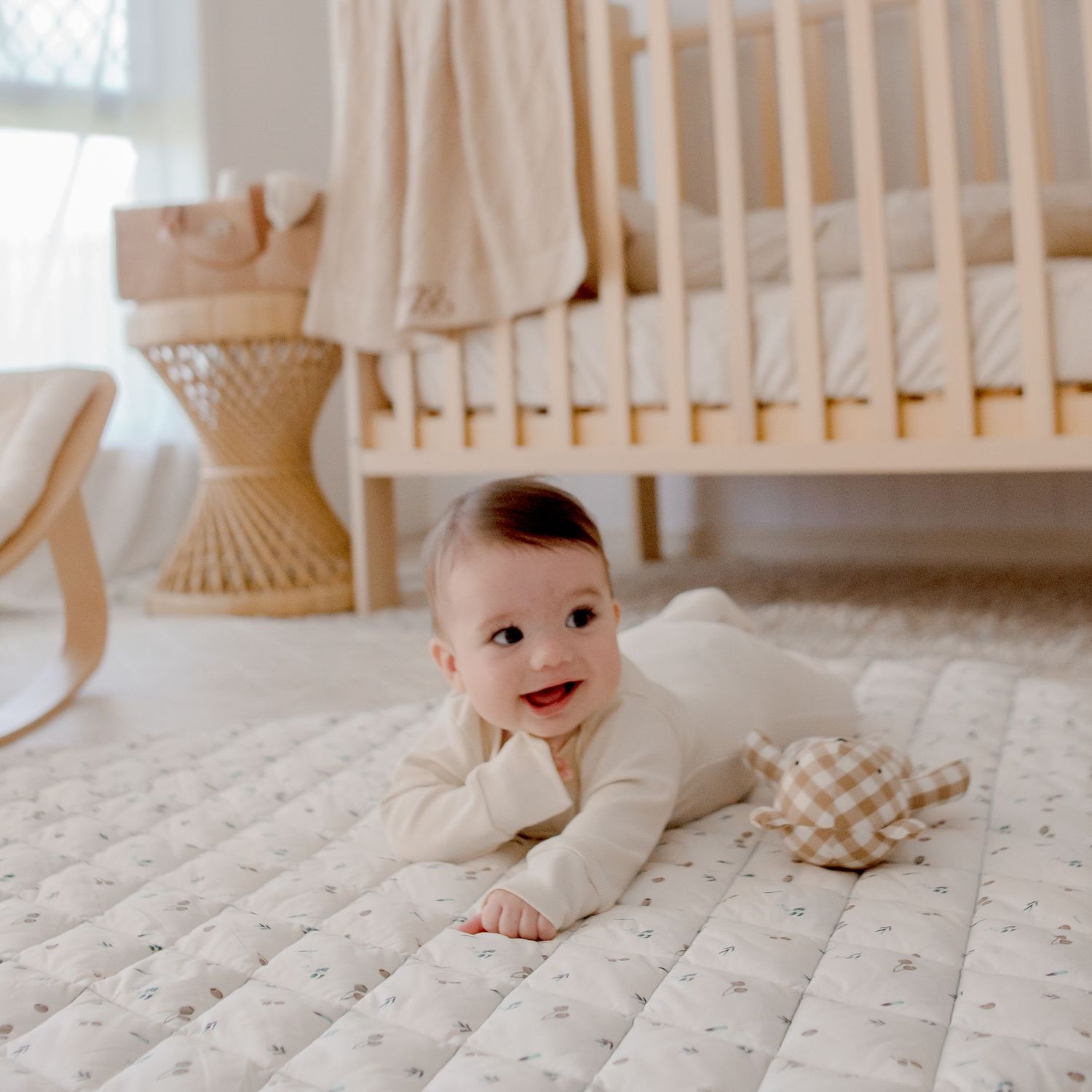 Baby in white onesie crawling on quilted mat beside plaid toy in neutral nursery.