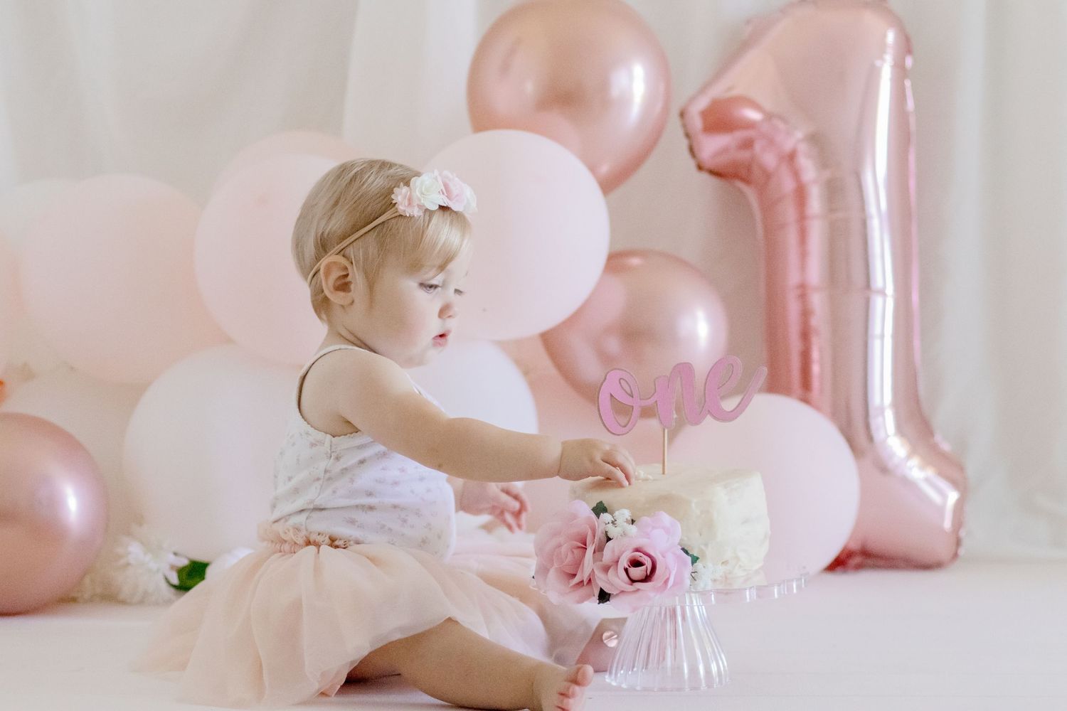 Baby in white dress sits among pink balloons with birthday cake at first birthday photoshoot.