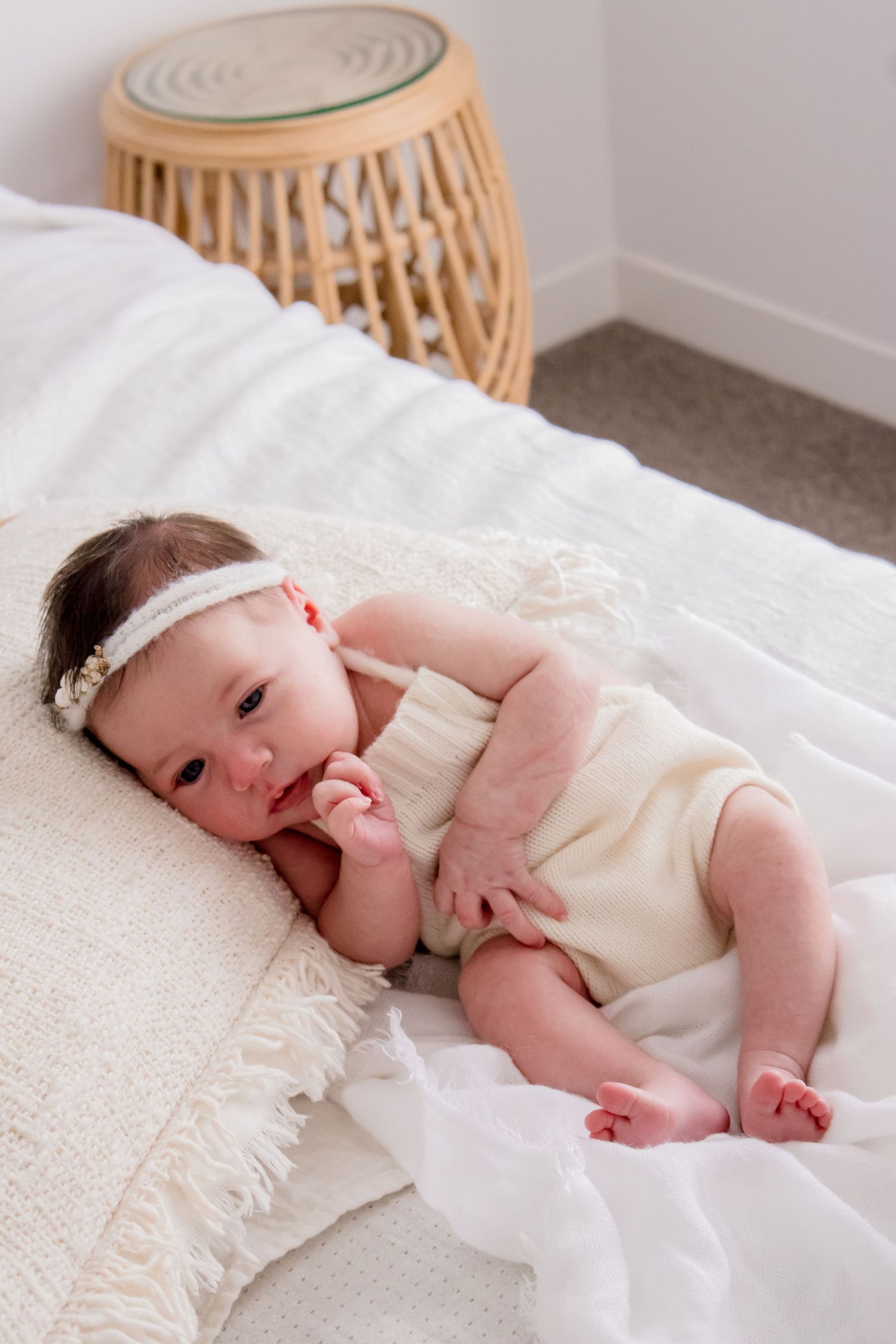 Baby in cream-colored outfit lying on white textured bedding near rattan furniture.
