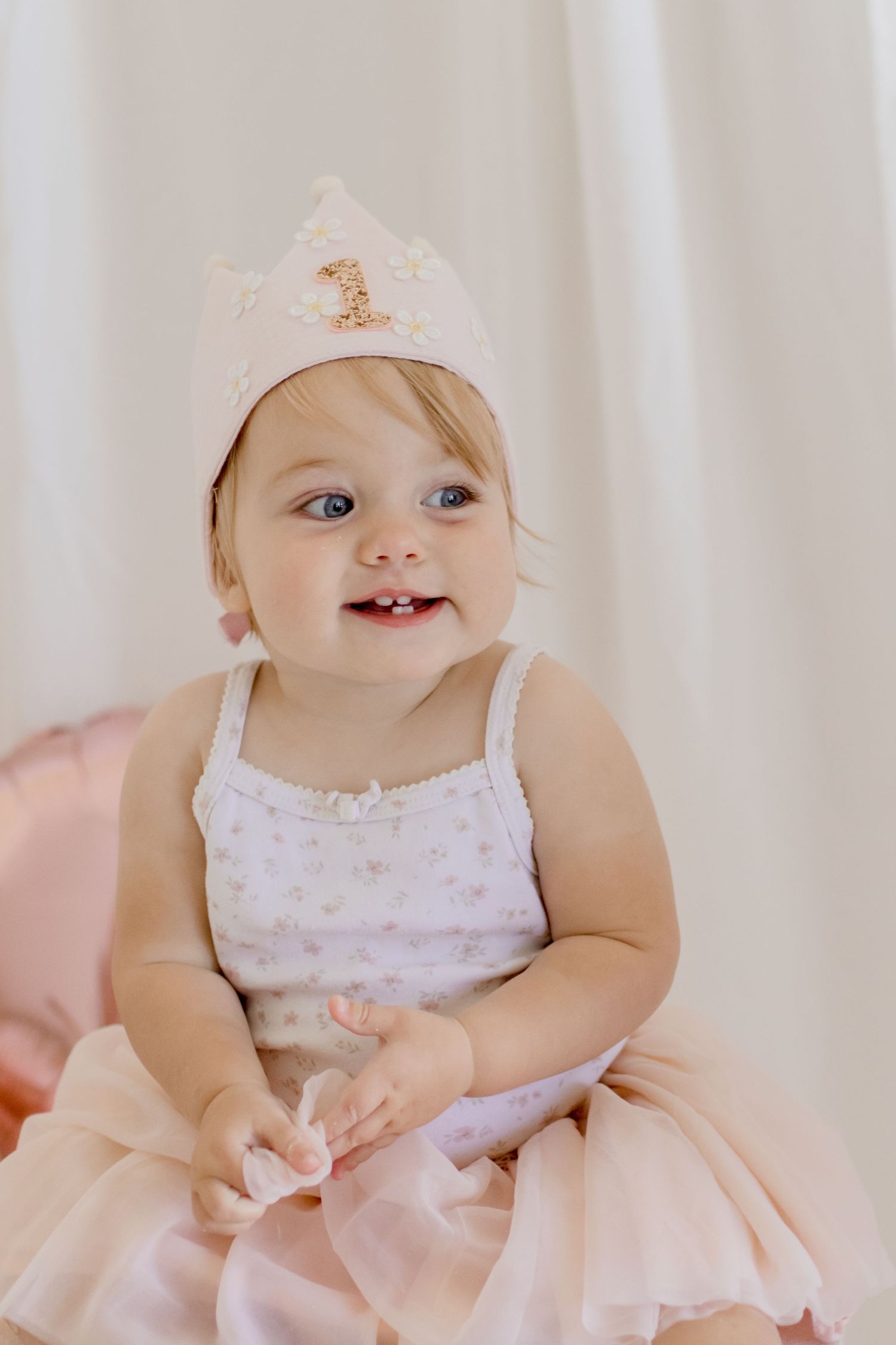 A joyful baby in pink tulle dress and crown smiles while sitting on white pedestal.
