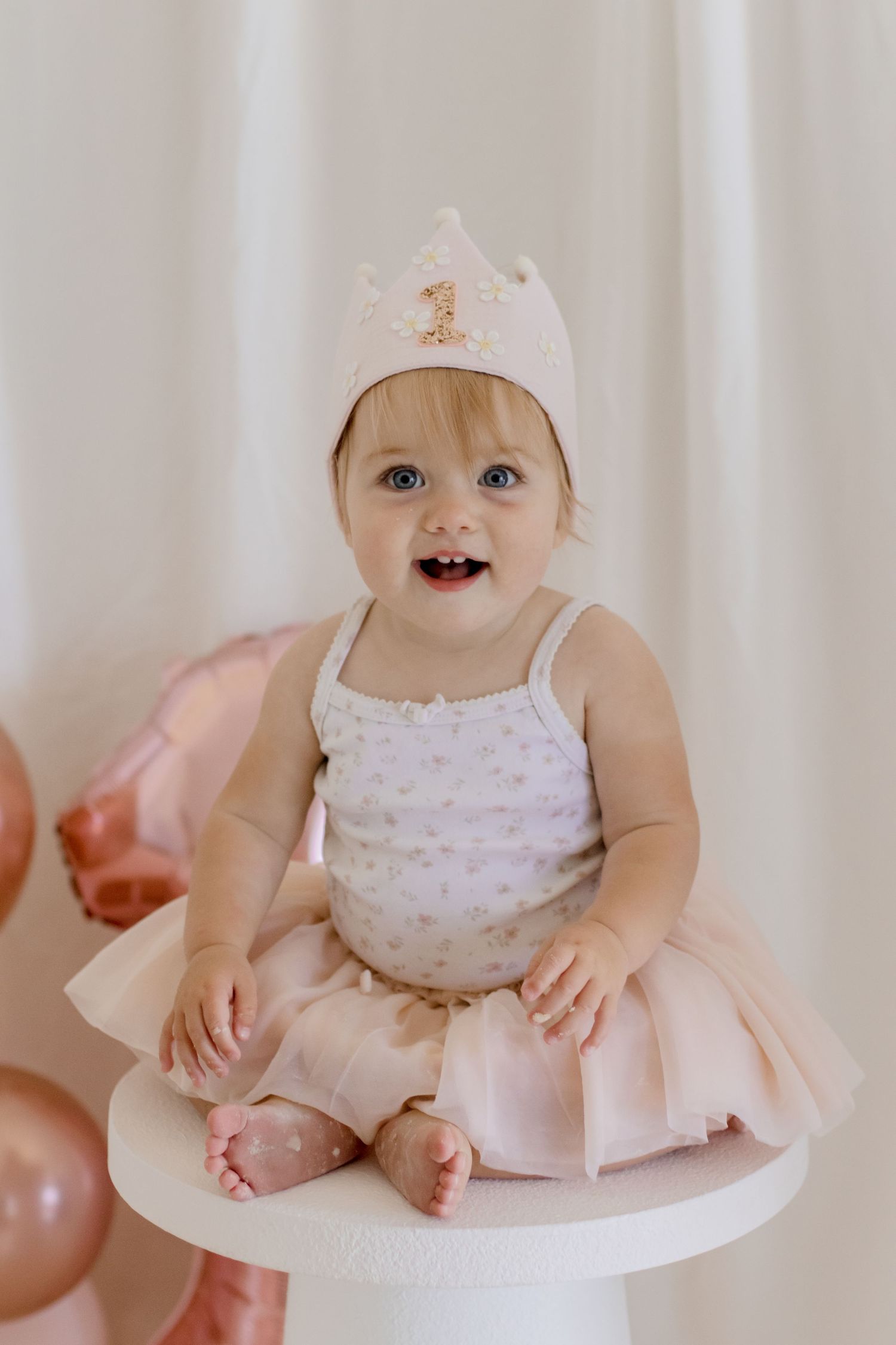 A smiling baby wearing a pink tutu and crown sits on a white pedestal with balloons in the background.